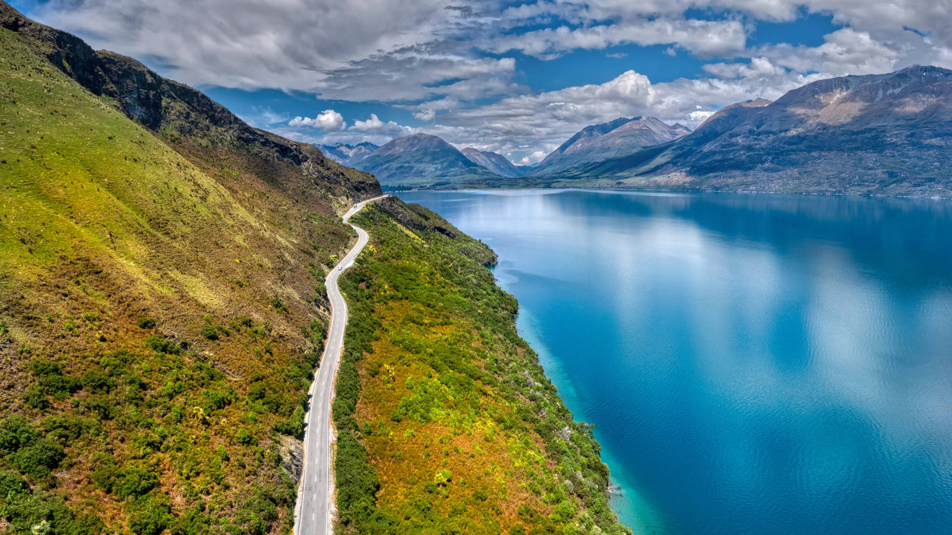 一條蜿蜒的道路沿著綠色山坡，旁邊是碧綠湖泊，背景有山脈。 — 風景動畫 壁紙