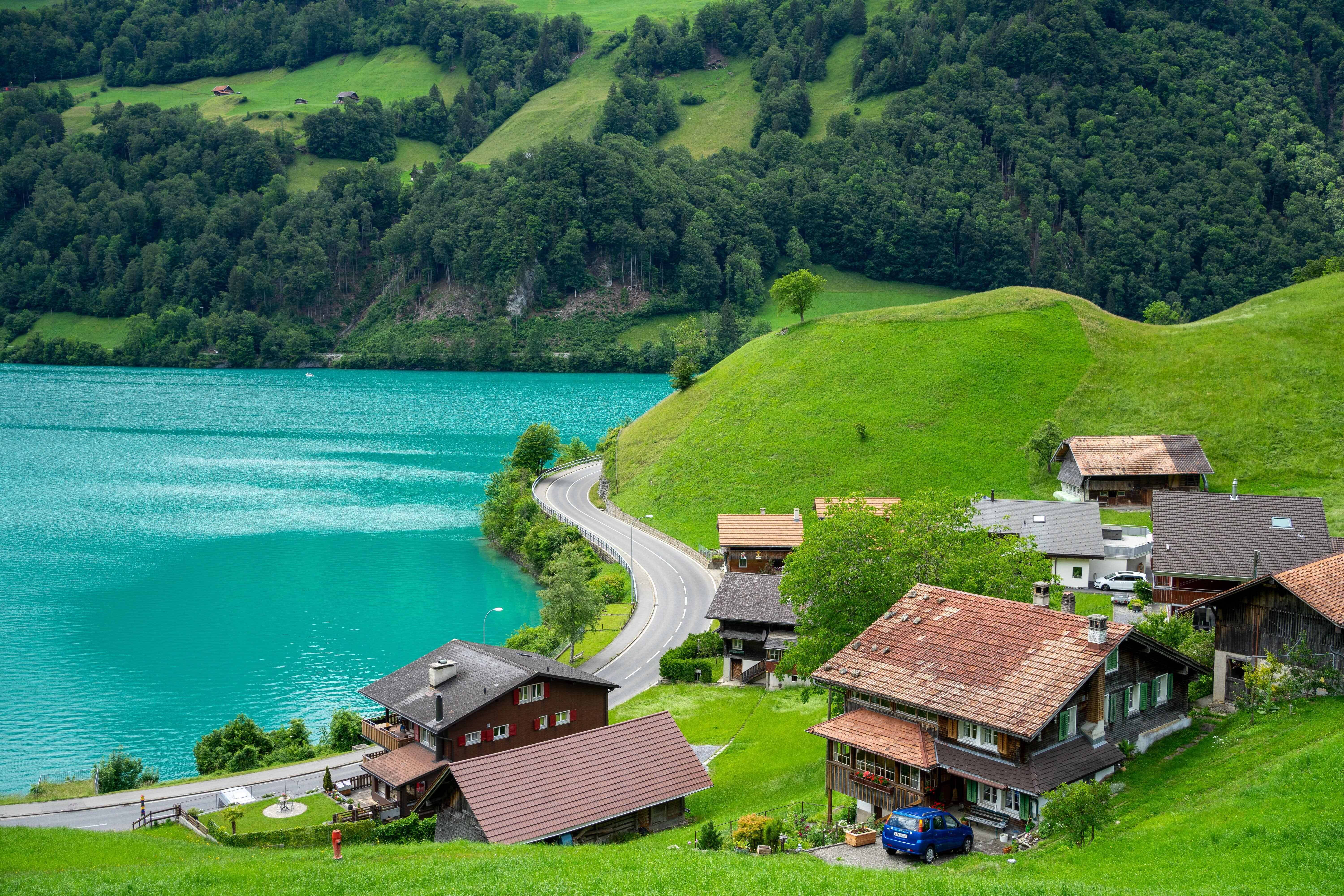 Lake 桌布 壁紙 — 一條蜿蜒的道路緊貼著充滿活力的綠松石湖，旁邊是坐落在鬱鬱蔥蔥的綠色山丘中的傳統木屋。