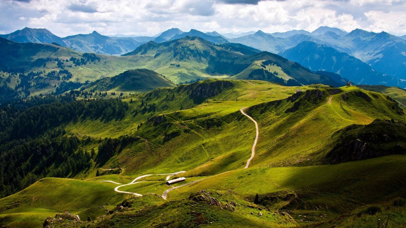山景 桌布 壁紙 — 一條蜿蜒的道路蜿蜒穿過充滿活力的綠色山谷，天空部分多雲。