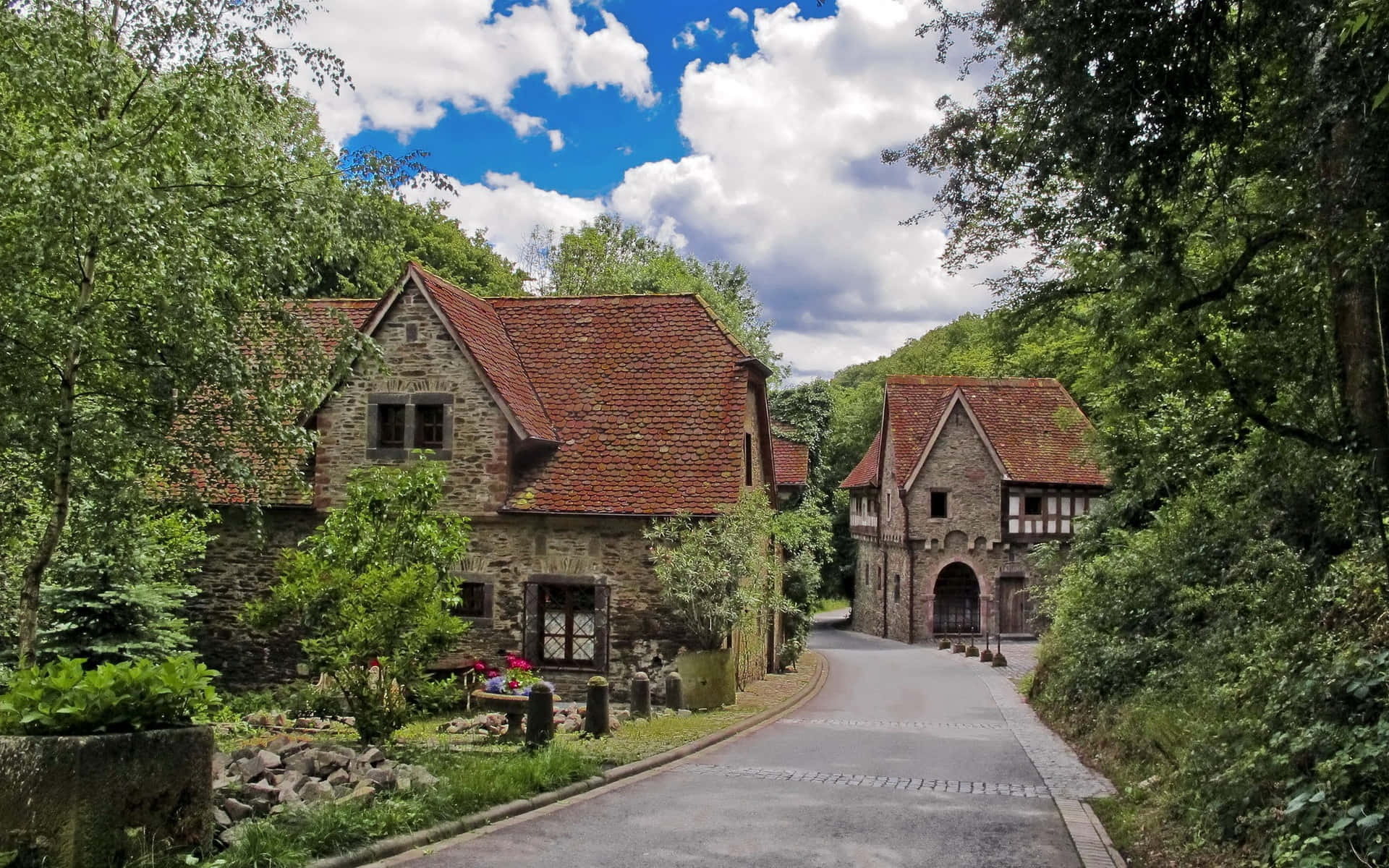 Village 桌布 壁紙 — 一條蜿蜒的道路蜿蜒經過石頭小屋，這些小屋擁有紅色瓦片屋頂，坐落在茂密的綠色森林中，上方是明亮的藍天和蓬鬆的雲朵。