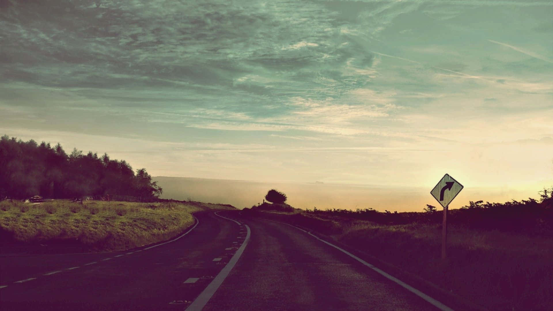 Road 桌布 壁紙 — 一條蜿蜒的鄉村道路在日落時分柔和的粉彩天空下蜿蜒伸向遠方，有一棵孤樹和方向標誌。