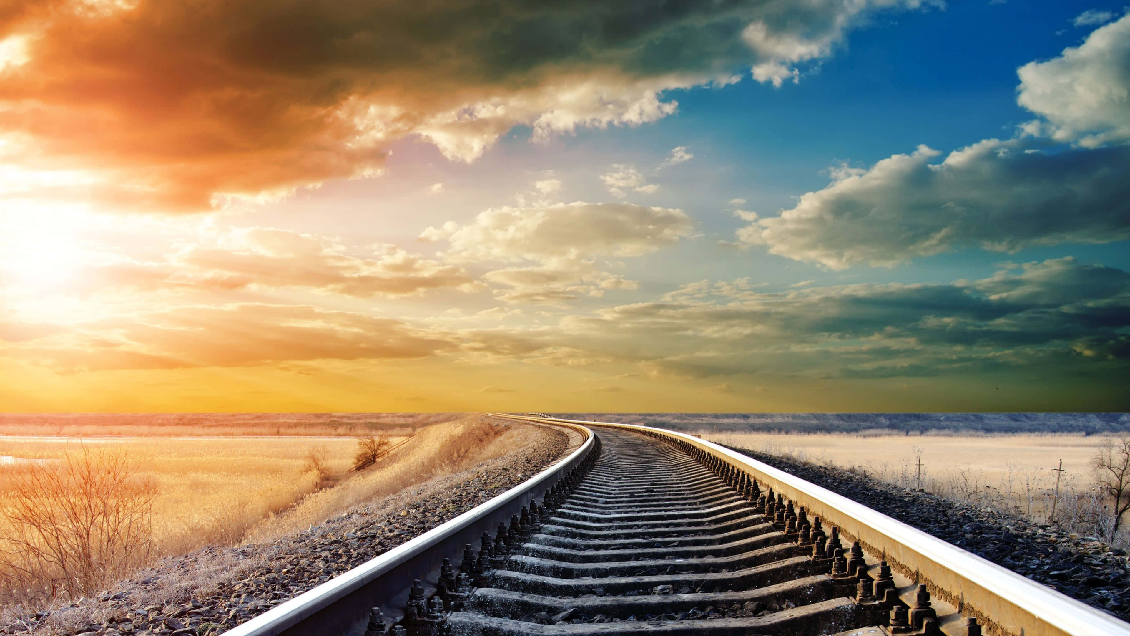 track 桌布 壁紙 — 一條蜿蜒的鐵路軌道在戲劇性的日落天空下，伴隨著金色的雲朵和結霜的田野，彎曲向地平線延伸。