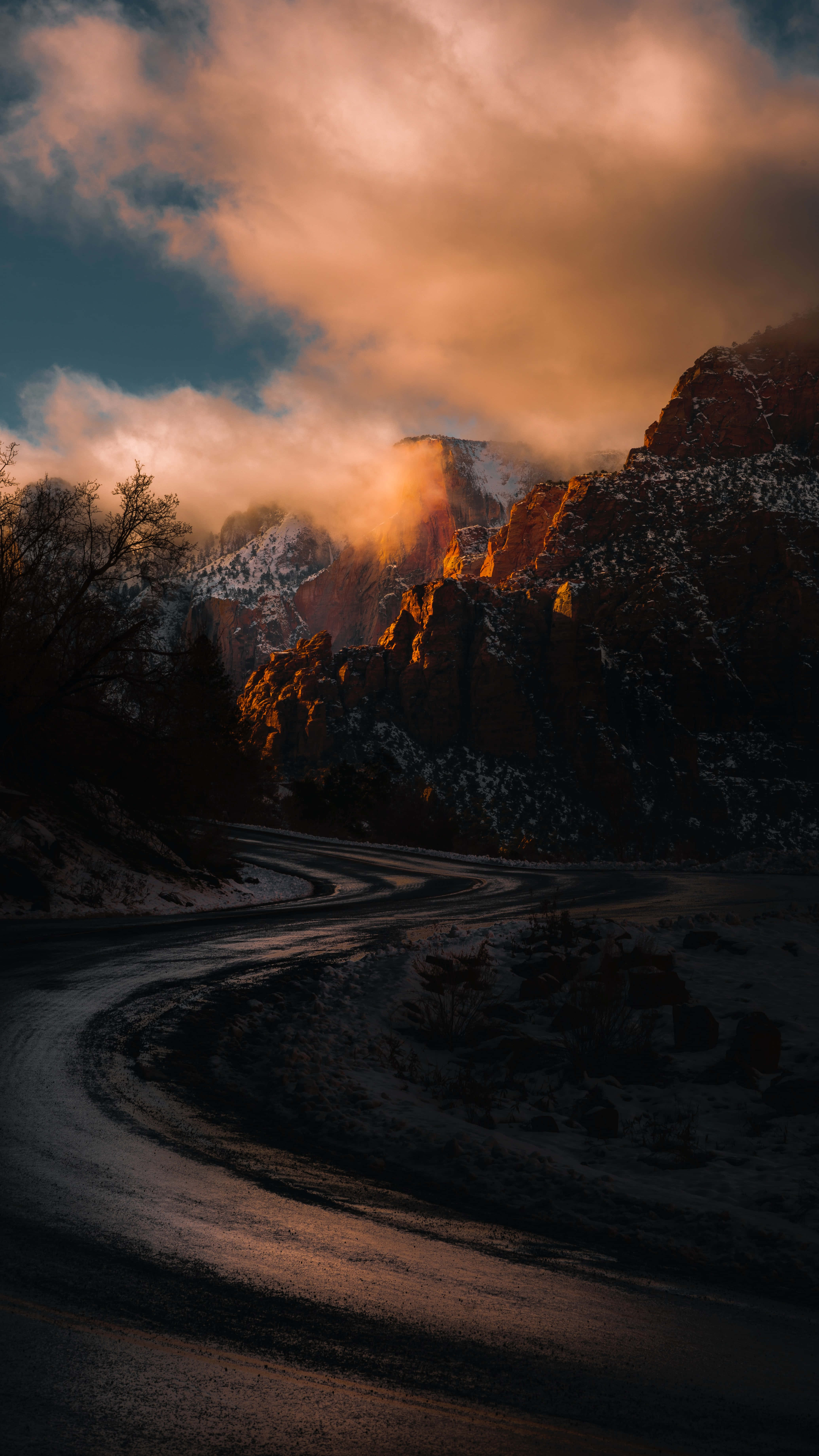 iPhone 桌布 壁紙 — 一條蜿蜒的雪路穿過戲劇性的山口，金色夕陽穿透雲層，照亮積雪的山峰。