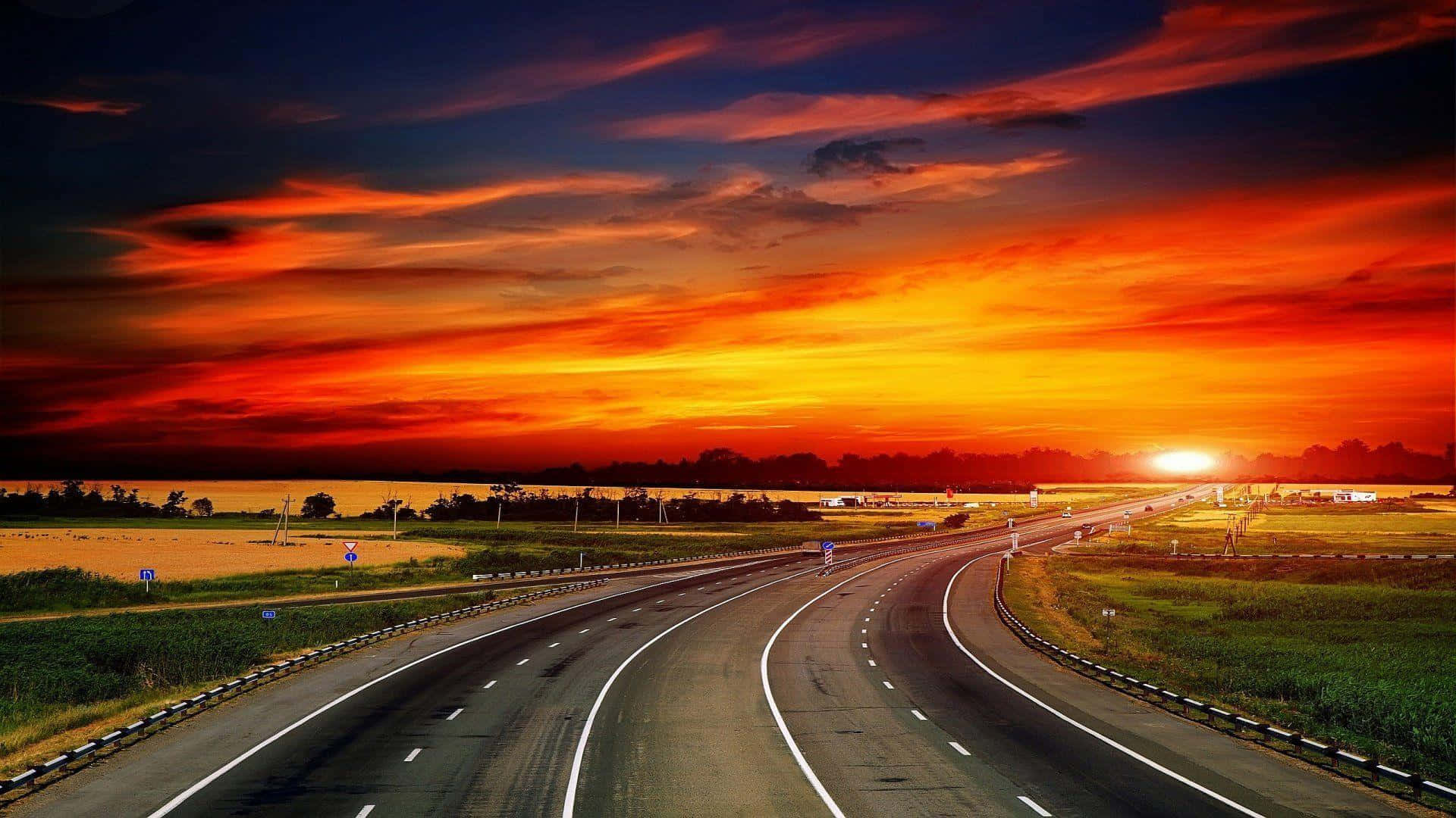 Highways 桌布 壁紙 — 一條蜿蜒的高速公路伸向熾熱的日落，開闊的田野上空有鮮豔的橙色、紅色和深藍色的雲彩。