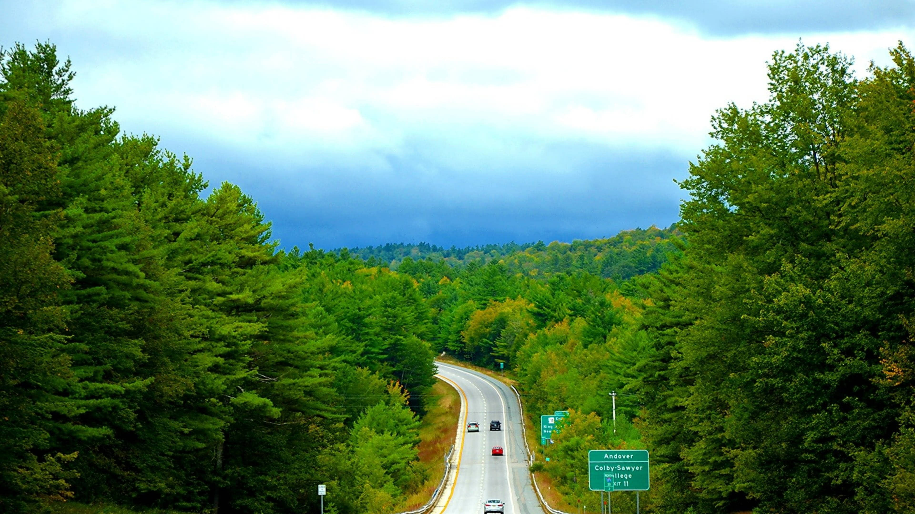 Vermont 桌布 壁紙 — 一條蜿蜒的高速公路穿過茂密的綠色森林，天空多雲，遠山壯麗。