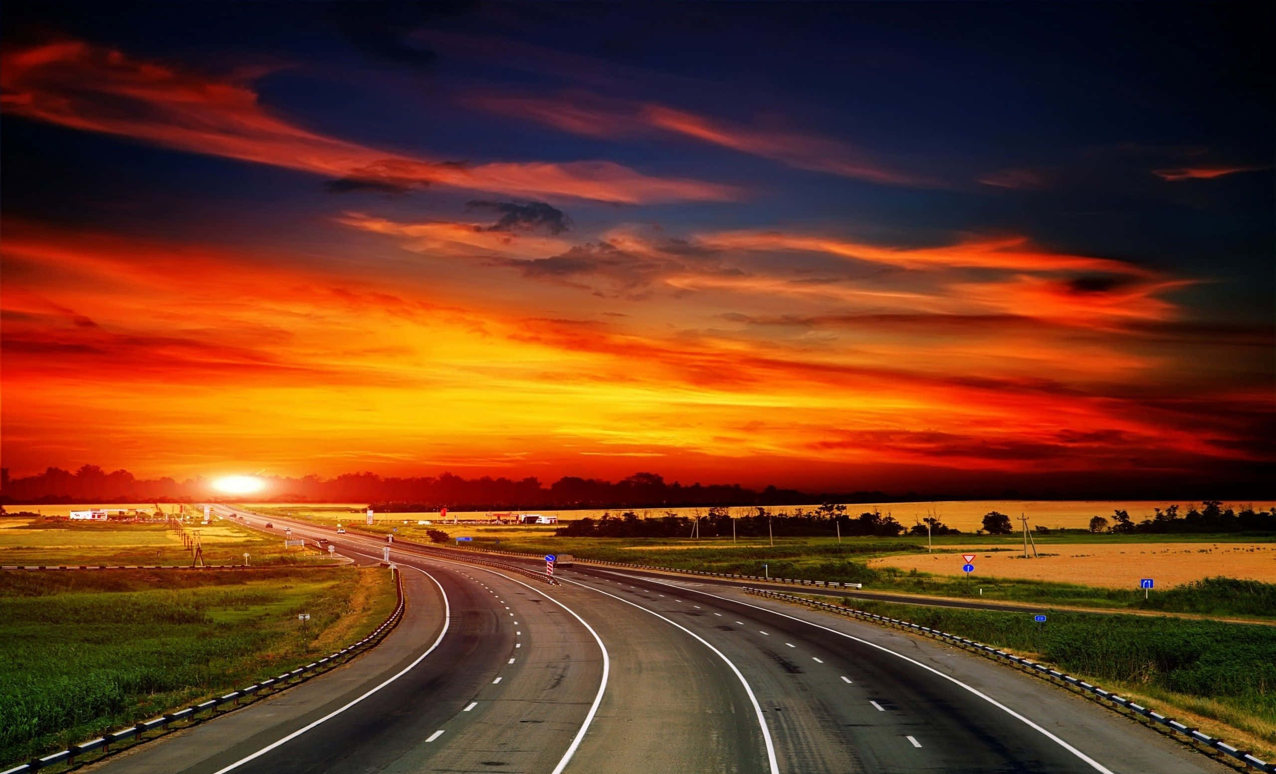highway 桌布 壁紙 — 一條蜿蜒的高速公路穿過金色田野，上方是火燒般的夕陽天空，帶有深橙色和紫色的雲彩。