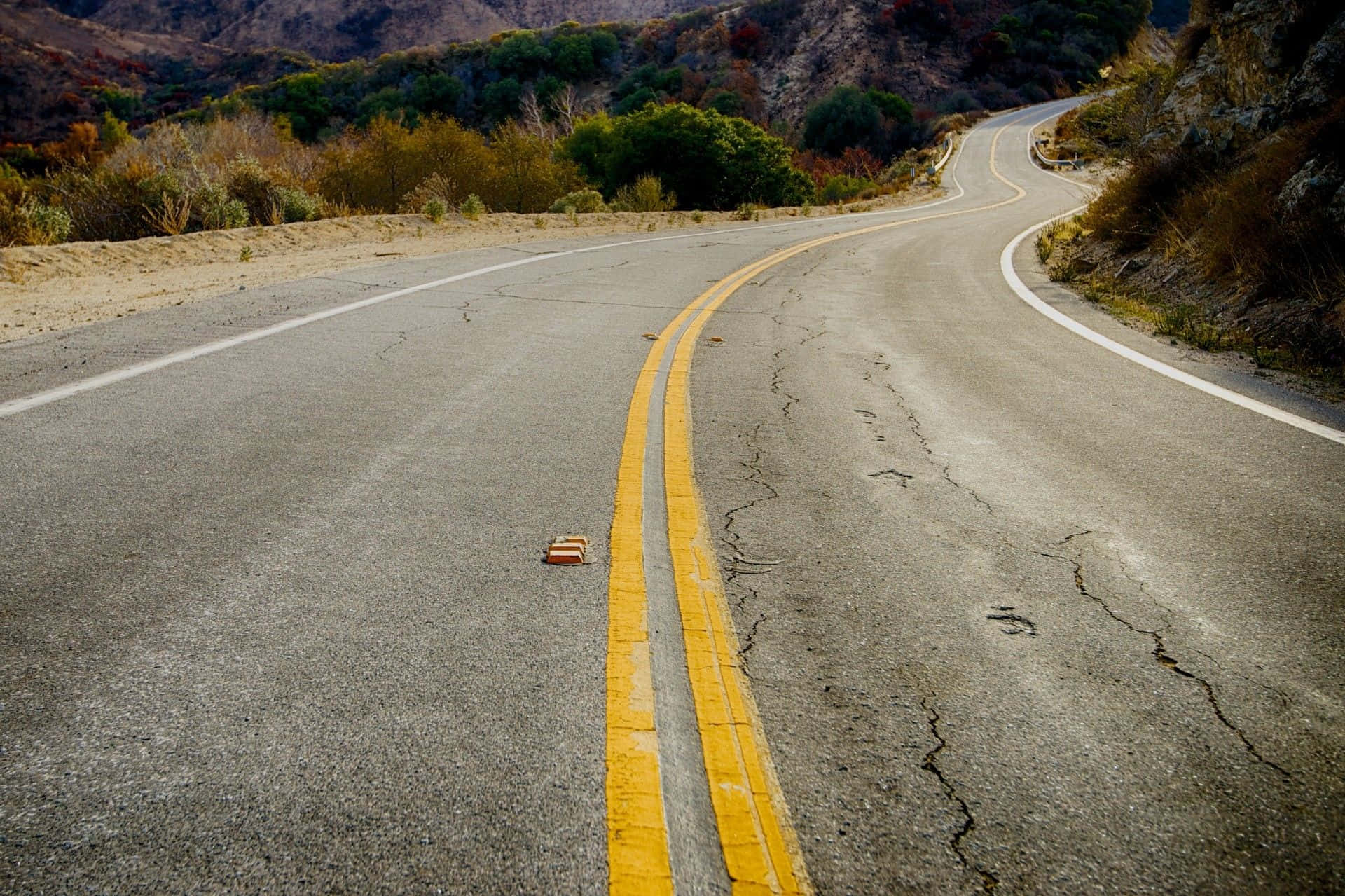 Road 桌布 壁紙 — 一條蜿蜒破裂的瀝青路在明亮的日光下穿過乾燥多山的地形，植被稀疏。