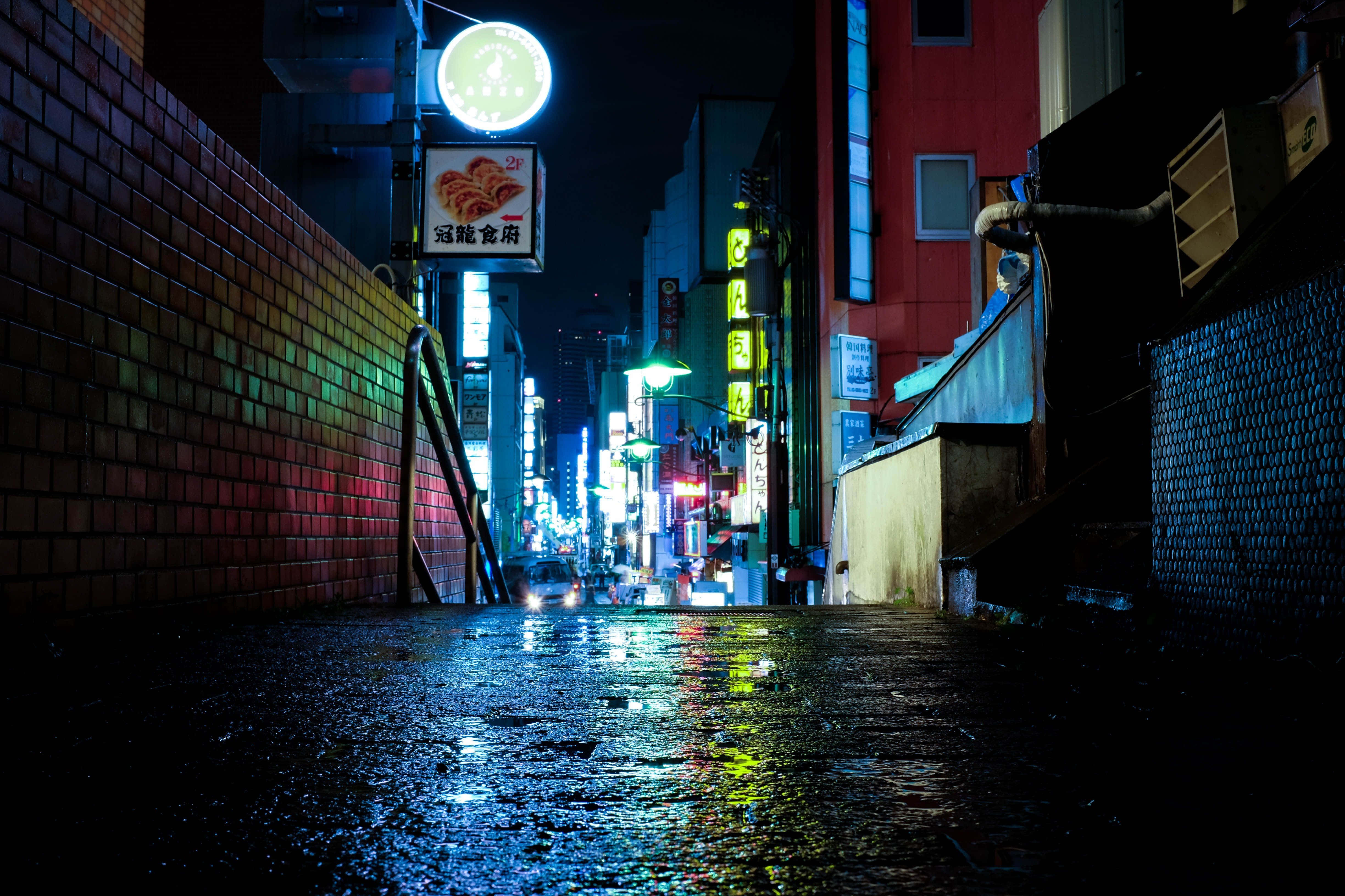 Alleyway 桌布 壁紙 — 一條被雨水打濕、霓虹燈閃爍的夜晚小巷，在黑暗的磚牆和建築物上反射出鮮豔的發光標誌。