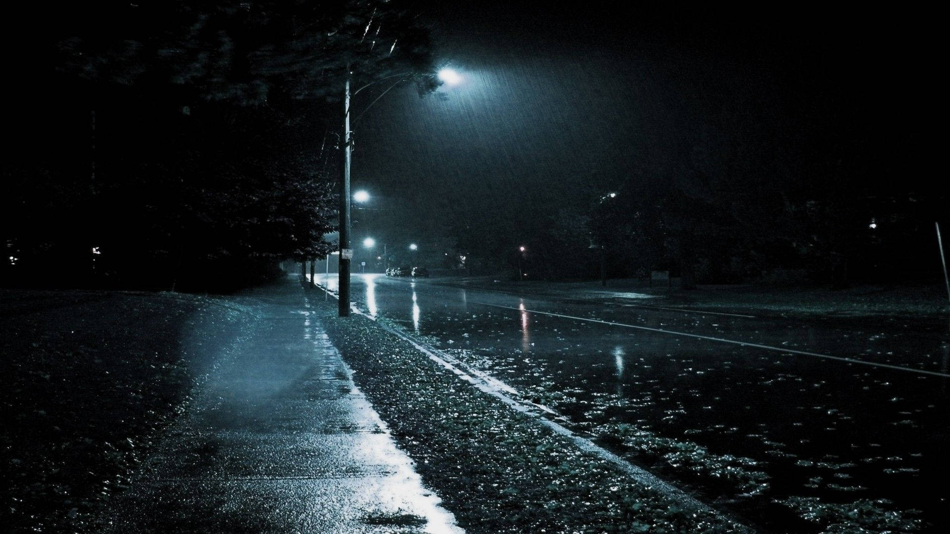 下雨 桌布 壁紙 — 一條被雨水打濕的夜間城市街道，被刺眼的街燈照亮，在落下的雨中投下長長的影子。
