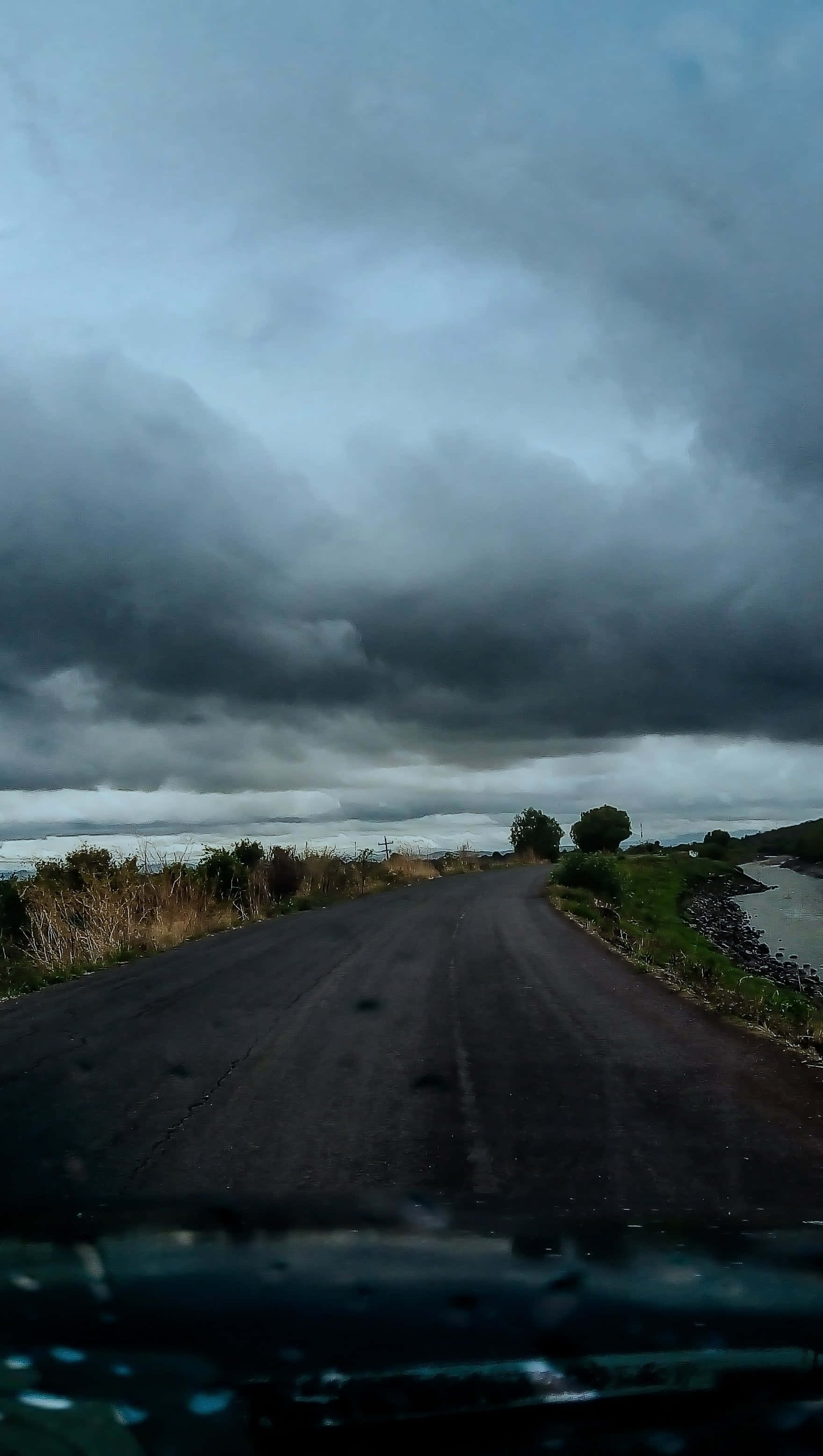 Weather 桌布 壁紙 — 一條被雨水打濕的道路蜿蜒伸向暴風雨的地平線，烏雲籠罩著遠處的水域。