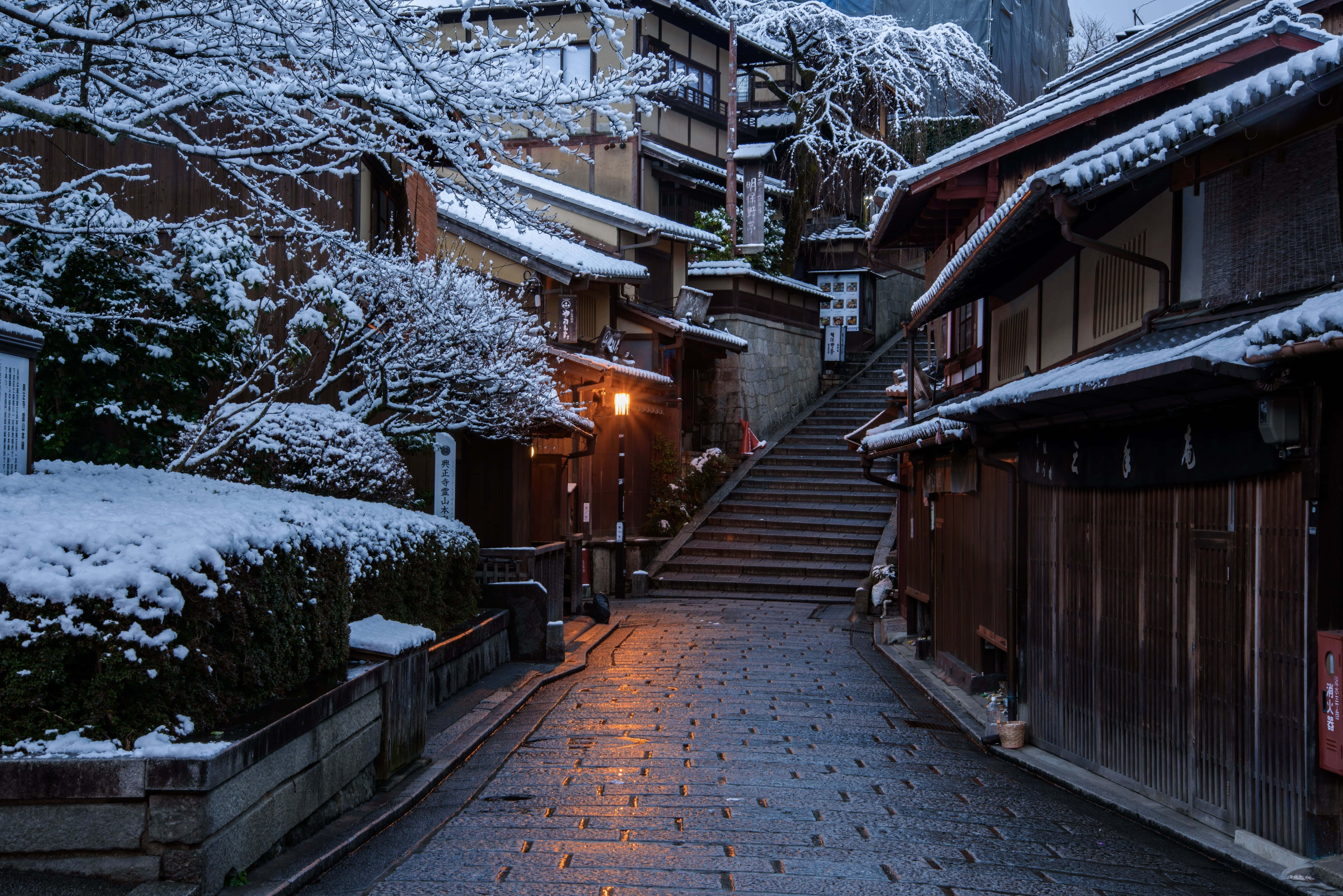 日本 桌布 壁紙 — 一條被雪覆蓋的傳統日本街道，夜晚溫暖的燈籠光映照在濕漉漉的石階上。
