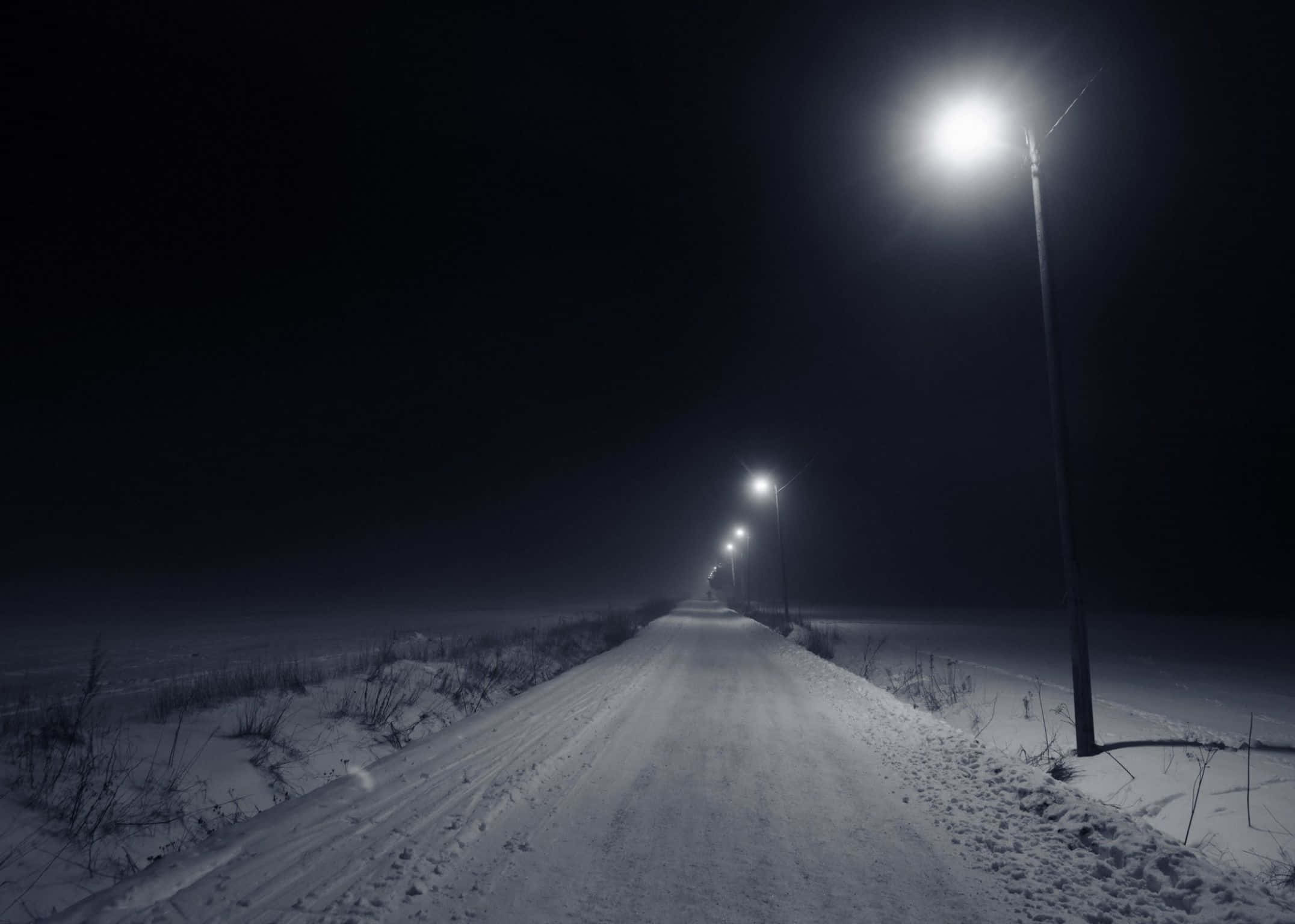 2152x1536 桌布 壁紙 — 一條被雪覆蓋的道路在寂靜的冬夜中，在一排發光街燈下延伸至黑暗之中。