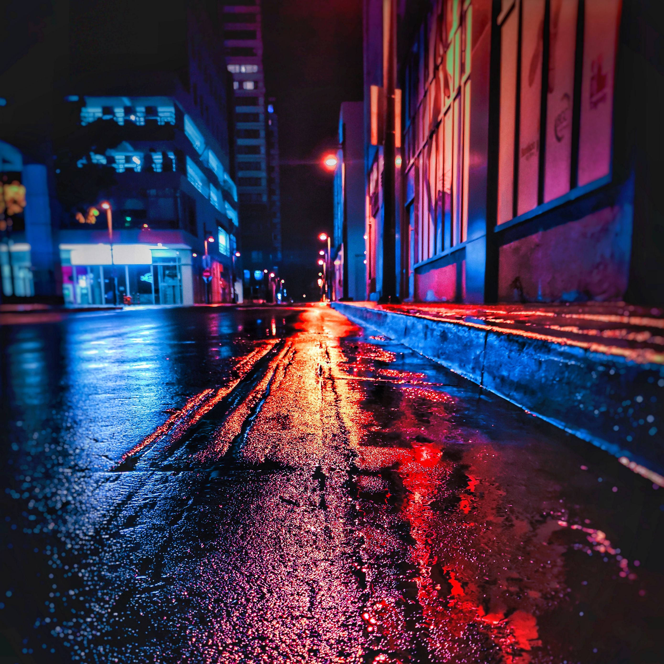城市 桌布 壁紙 — 一條被霓虹燈浸透的城市街道在夜晚將鮮豔的紅色和藍色燈光反射在濕漉漉的路面上，周圍是高聳的建築物。