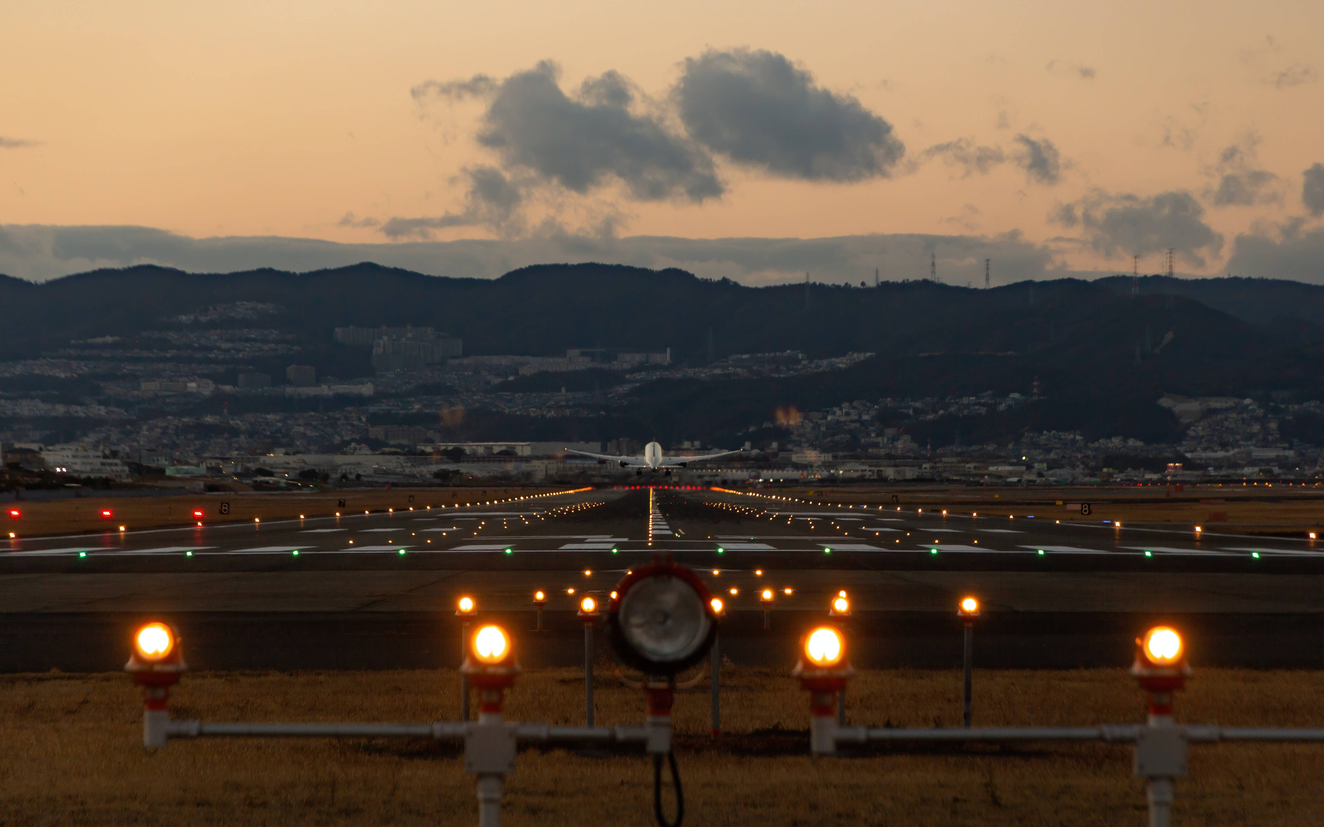 Runway 桌布 壁紙 — 一條跑道在黃昏時分延伸向遠處的飛機，背後有發光的燈光和剪影般的山丘。