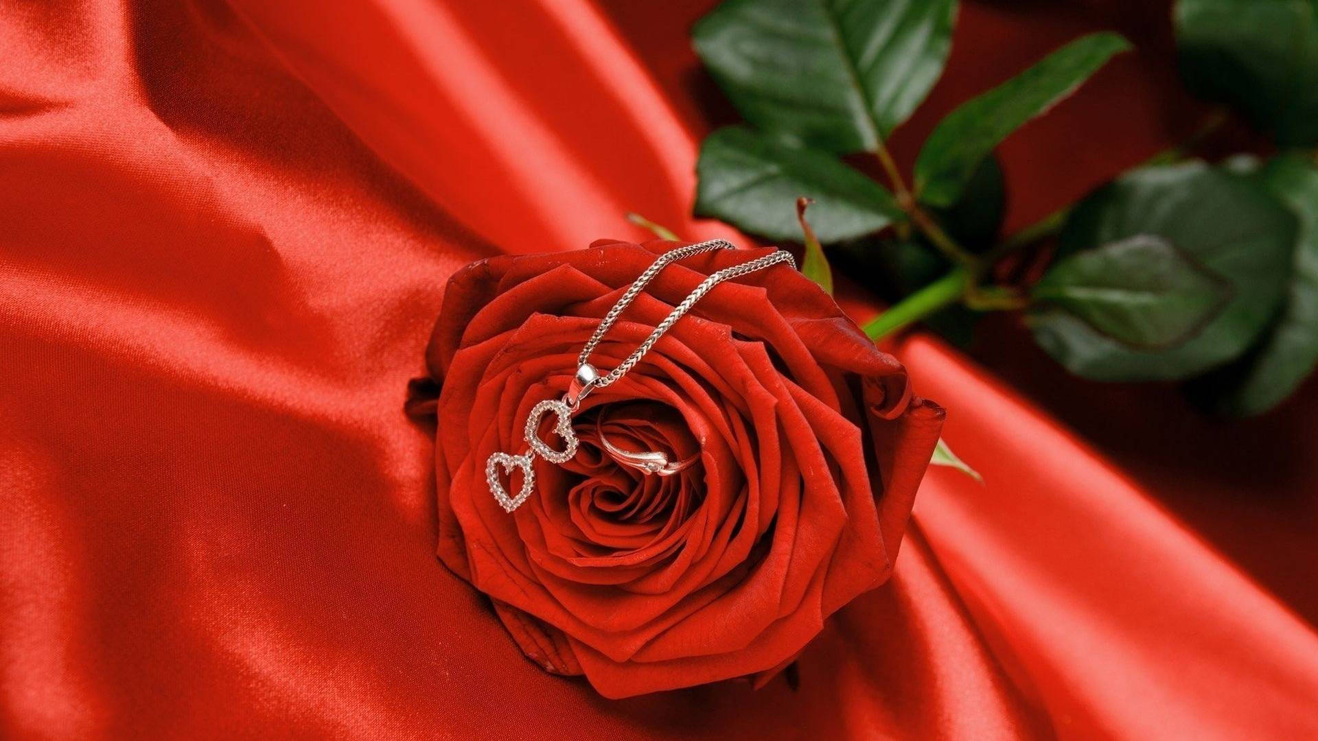 Red Roses 桌布 壁紙 — 一條銀色心形項鍊優雅地放在一朵鮮紅的玫瑰上，背景是豪華的紅色緞面。