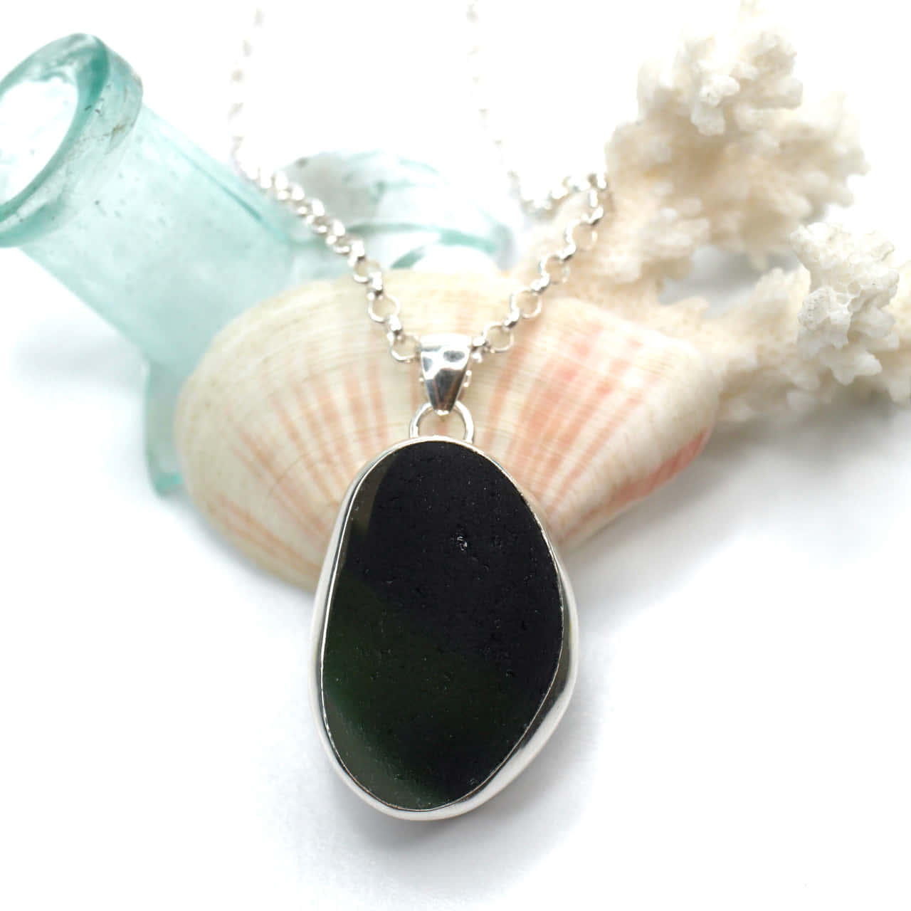 Sea Glass 桌布 壁紙 — 一條銀鏈上的時尚黑色海玻璃吊墜，旁邊放著貝殼和珊瑚，背景為亮白色。