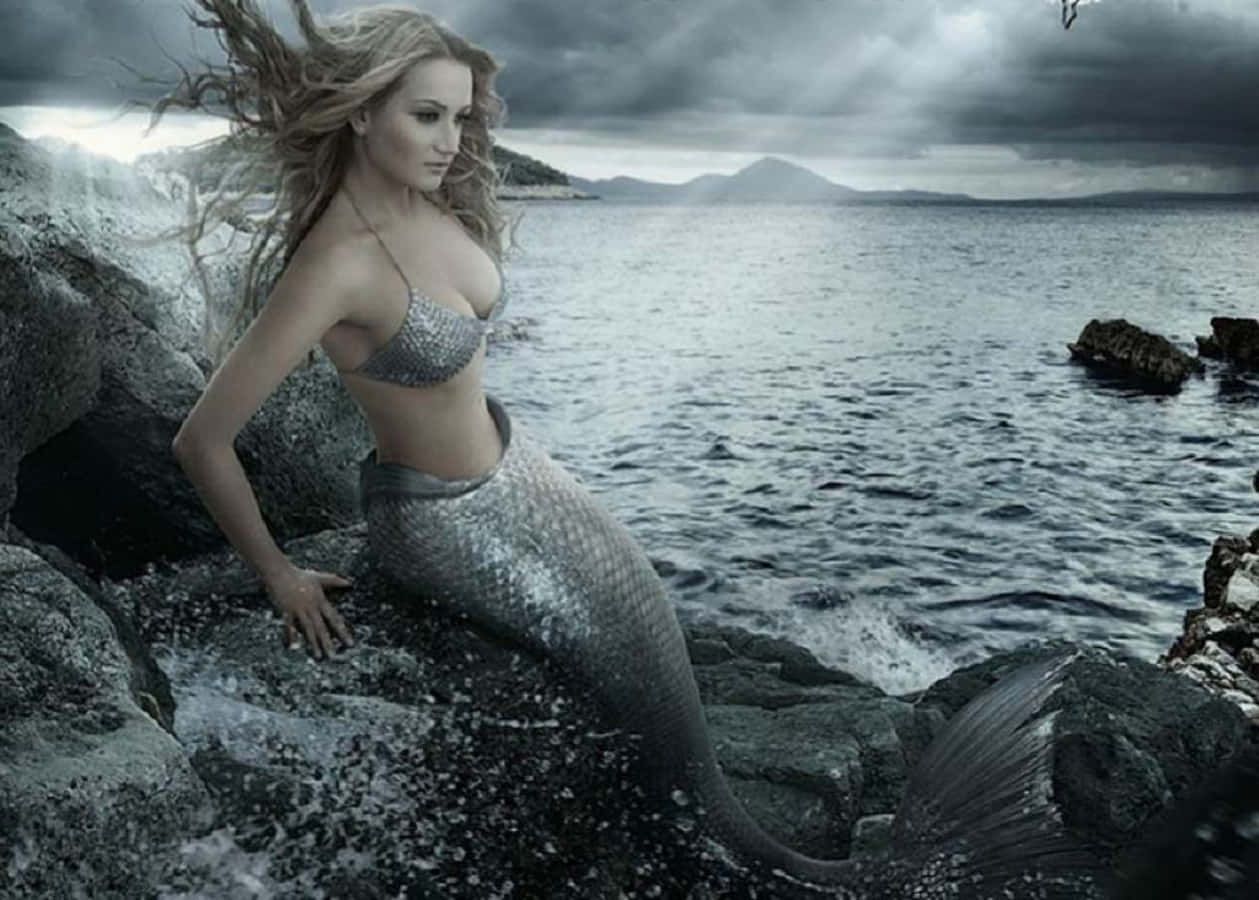 Mermaids 桌布 壁紙 — 一條銀鱗金髮飄逸的美人魚棲息在風暴中的海岸岩石上，在陰暗多雲的天空下若有所思地凝視著波濤洶湧的大海。
