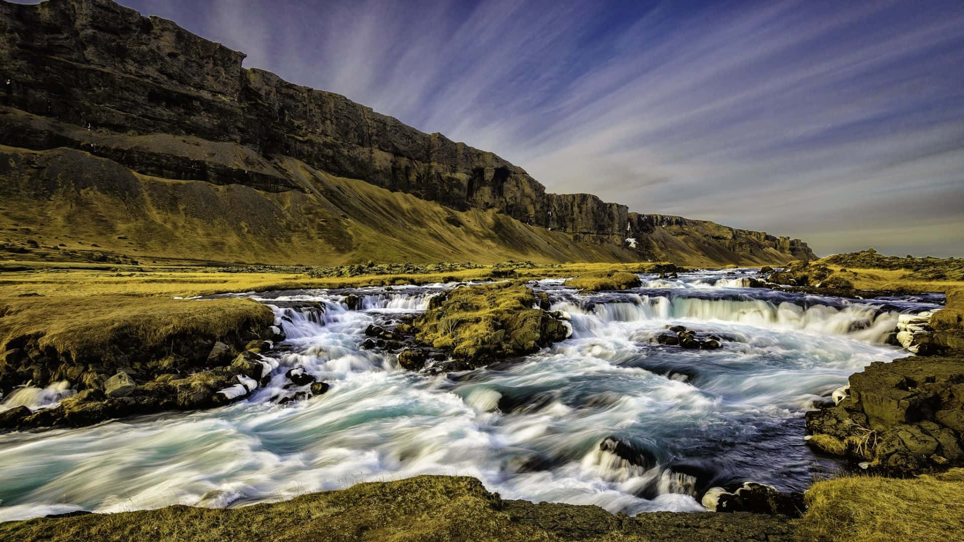 Iceland Desktop 桌布 壁紙 — 一條長時間曝光的河流在條紋天空下，衝過長滿苔蘚的岩石，朝向高聳的懸崖。