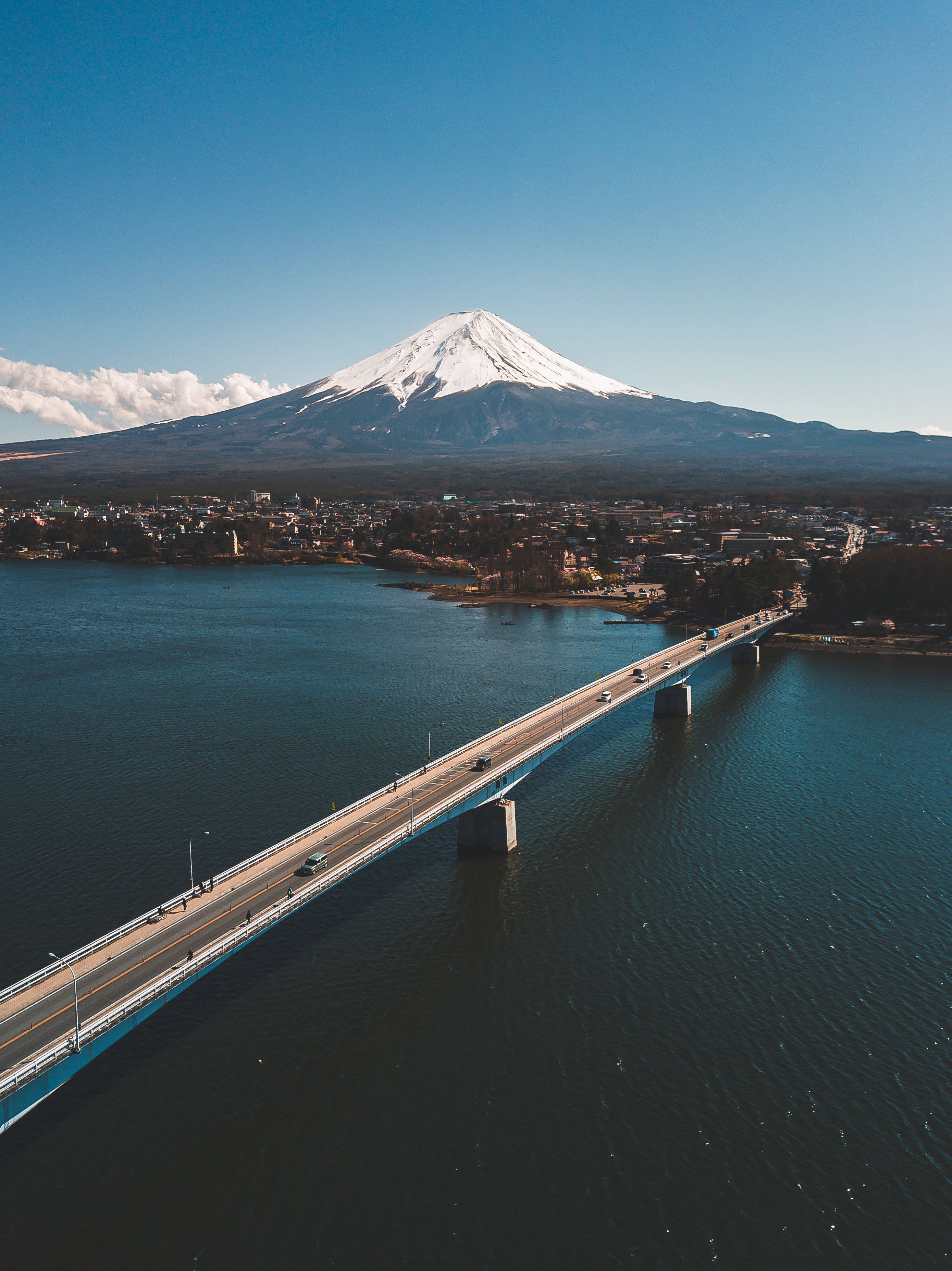 富士山 桌布 壁紙 — 一條長橋橫跨深藍色湖泊的航拍圖，背景中白雪覆蓋的富士山在清澈的藍天下雄偉地聳立。
