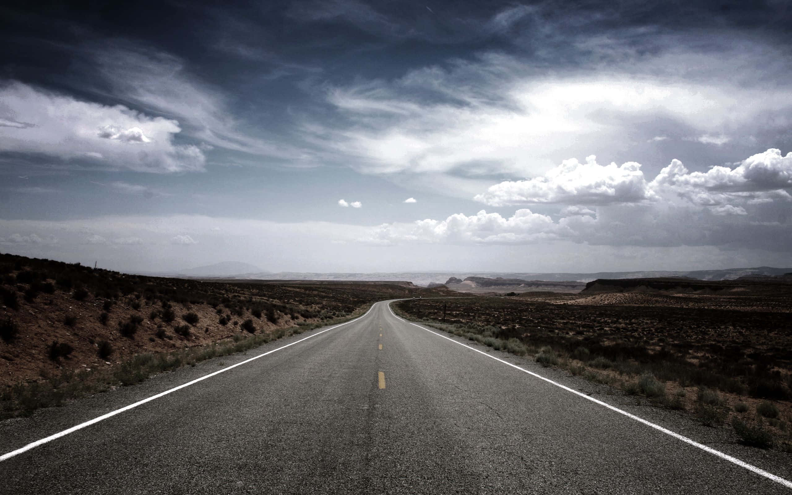 Road 桌布 壁紙 — 一條長直瀝青路在戲劇性的多雲天空下延伸至廣闊的沙漠。