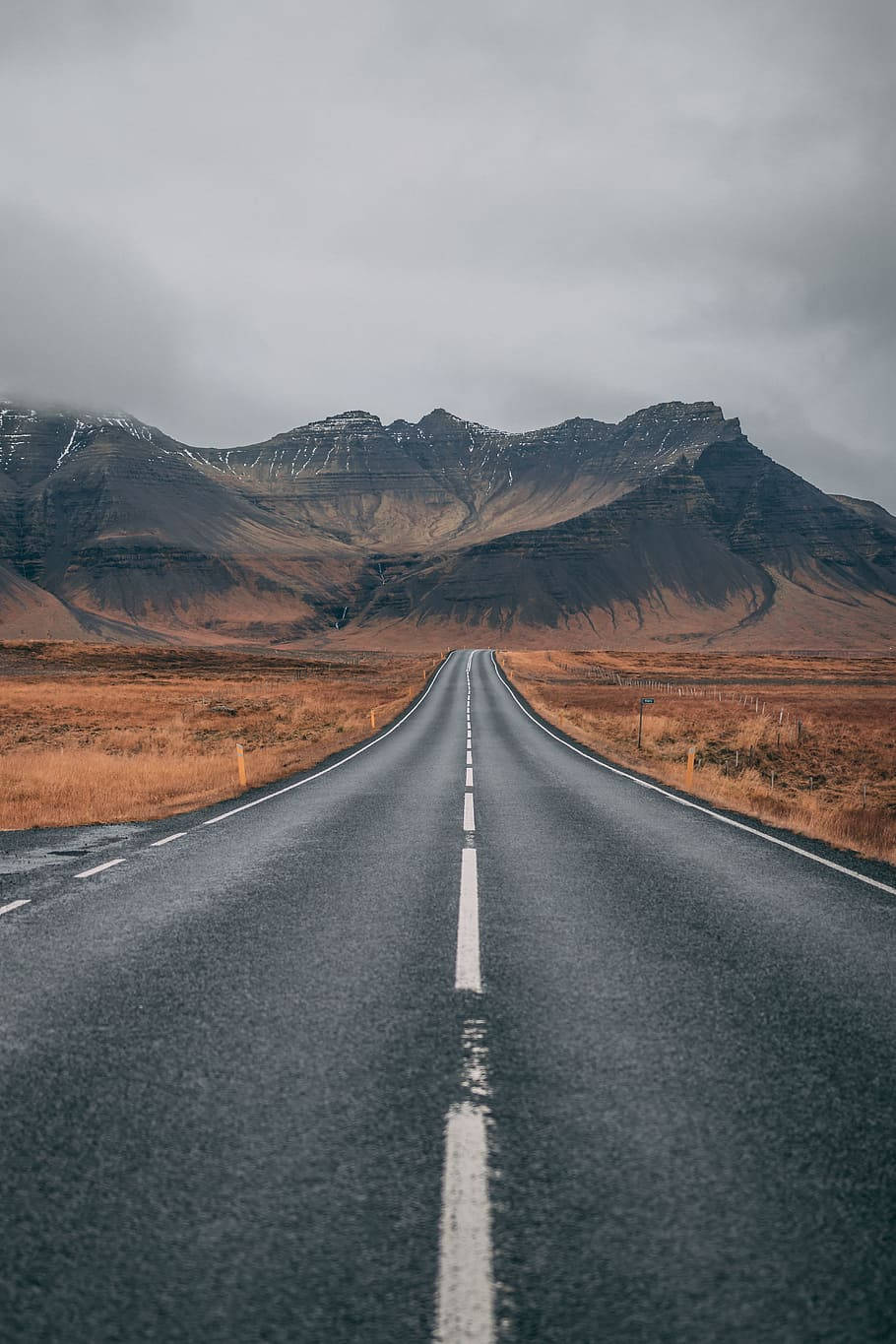 Road 桌布 壁紙 — 一條長直的道路延伸向遠方的山脈，天空多雲，兩旁有乾草。