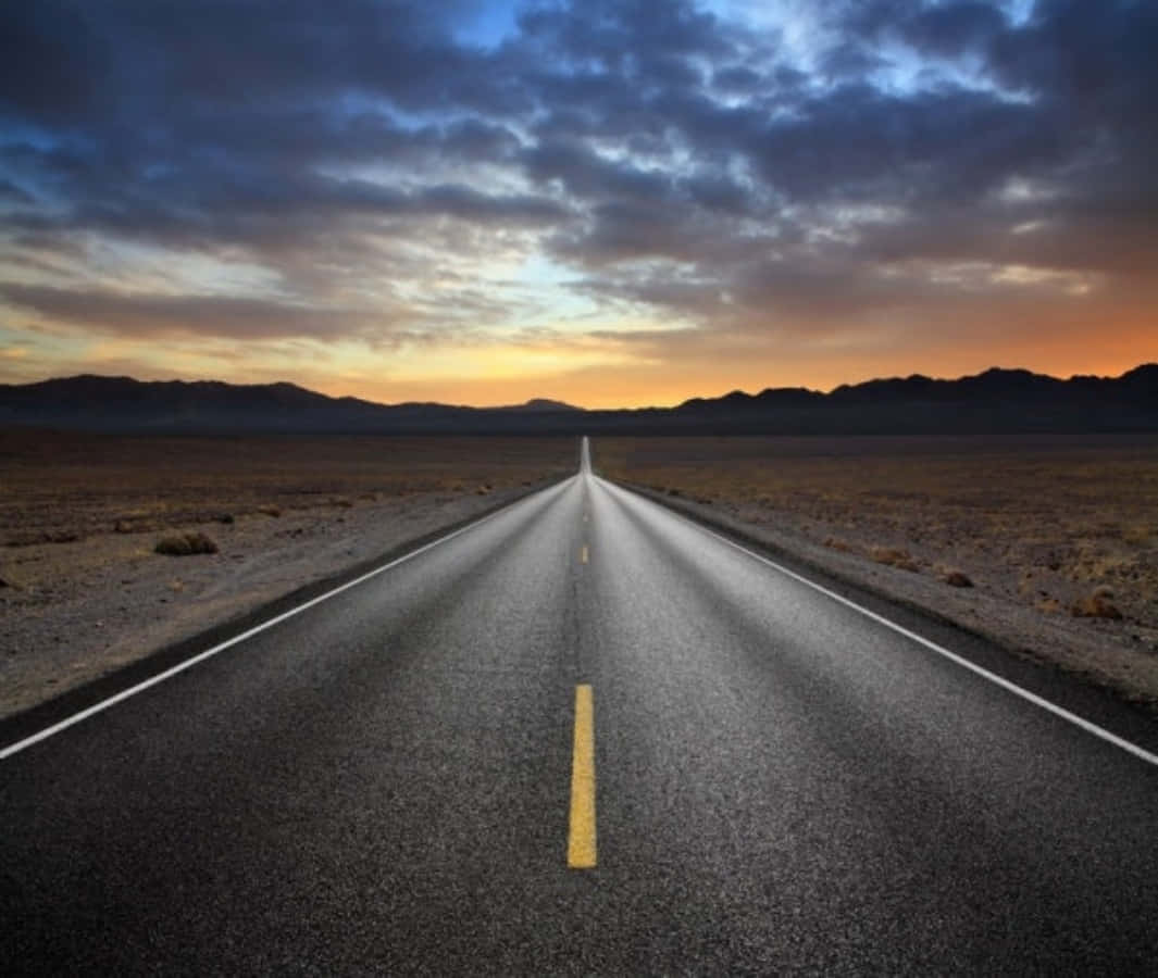 Highways 桌布 壁紙 — 一條長直的道路穿過貧瘠的沙漠，在戲劇性的暮色天空下延伸至地平線。