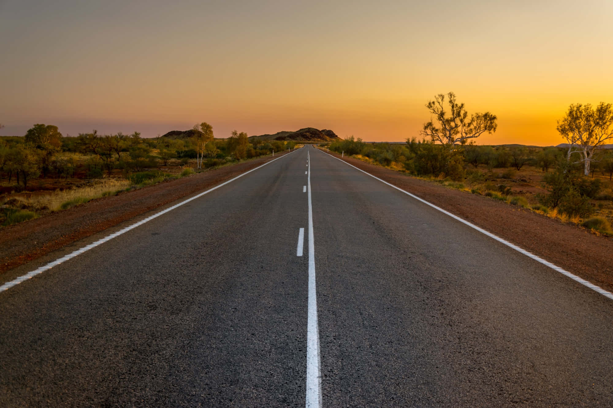 highway 桌布 壁紙 — 一條長直路在溫暖的日落天空下延伸至地平線，周圍是乾旱的景觀。