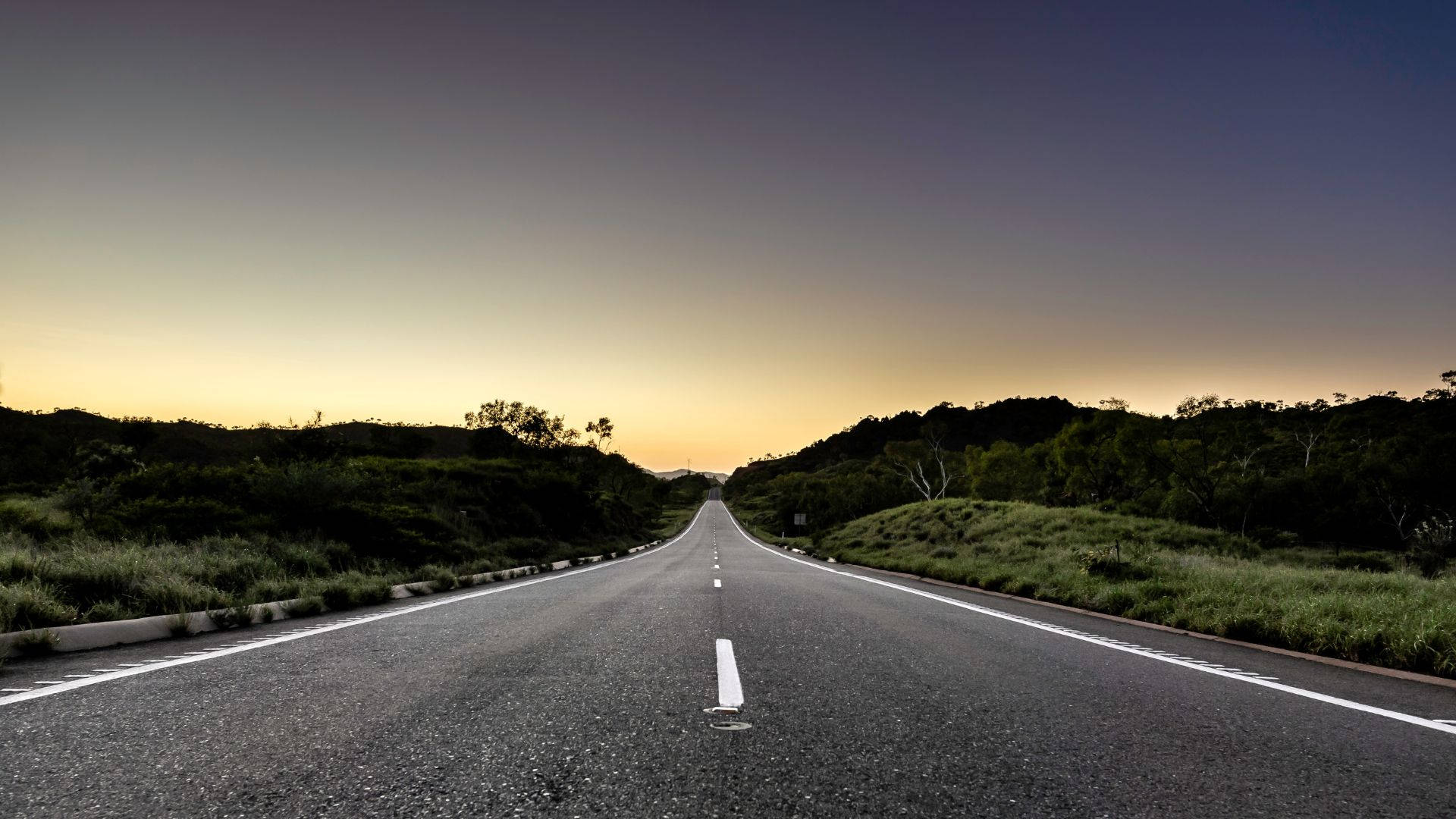 Road 桌布 壁紙 — 一條長直路在漸變日落天空下延伸至地平線，兩旁是綠色的山丘和樹木。