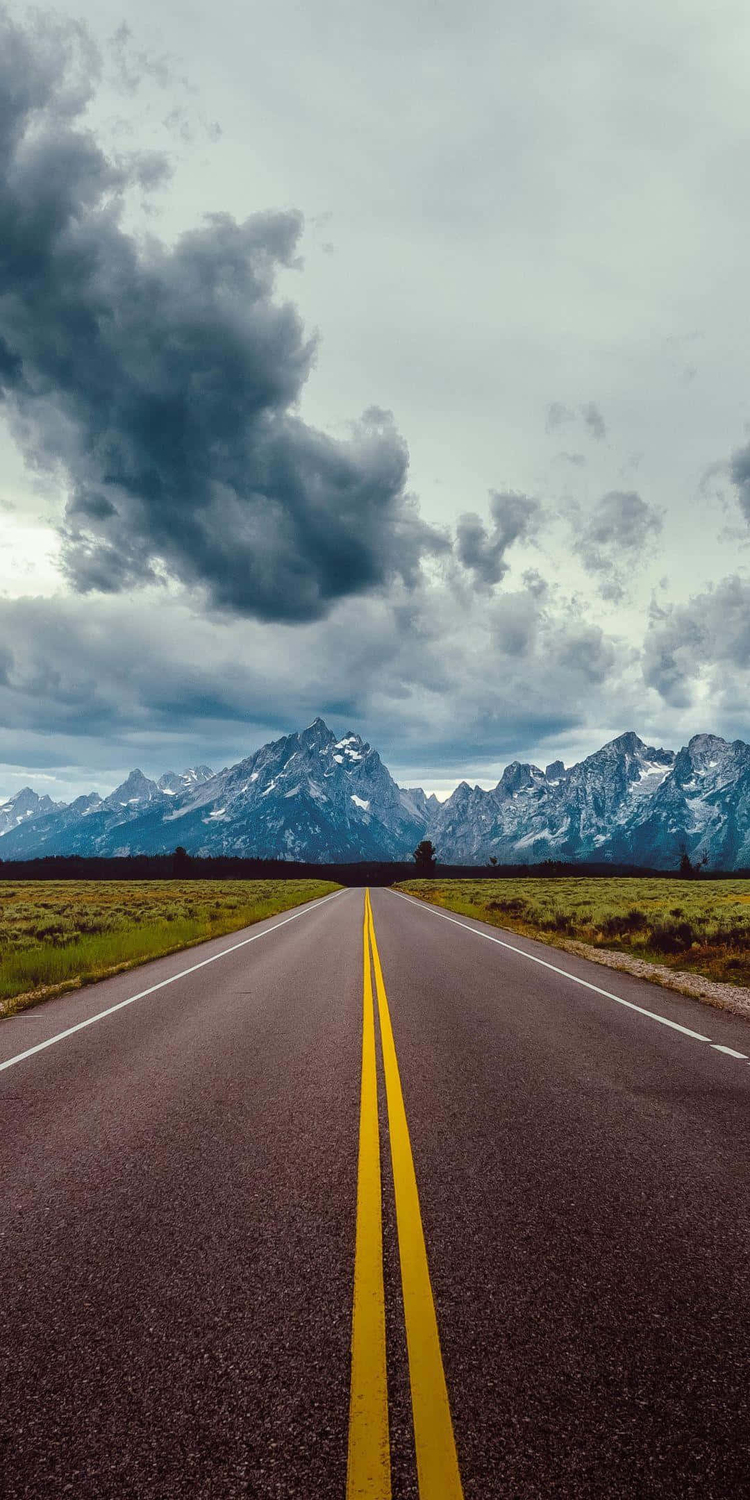 Highways 桌布 壁紙 — 一條長直路帶有黃線，在戲劇性的多雲天空下延伸至積雪覆蓋的山脈。