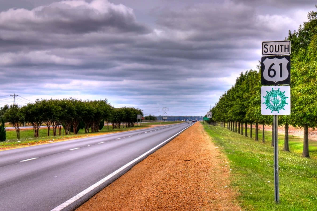 highway 桌布 壁紙 — 一條長直高速公路在戲劇性的多雲天空下延伸，兩旁有綠樹和碎石路肩，並有一個South 61路標。