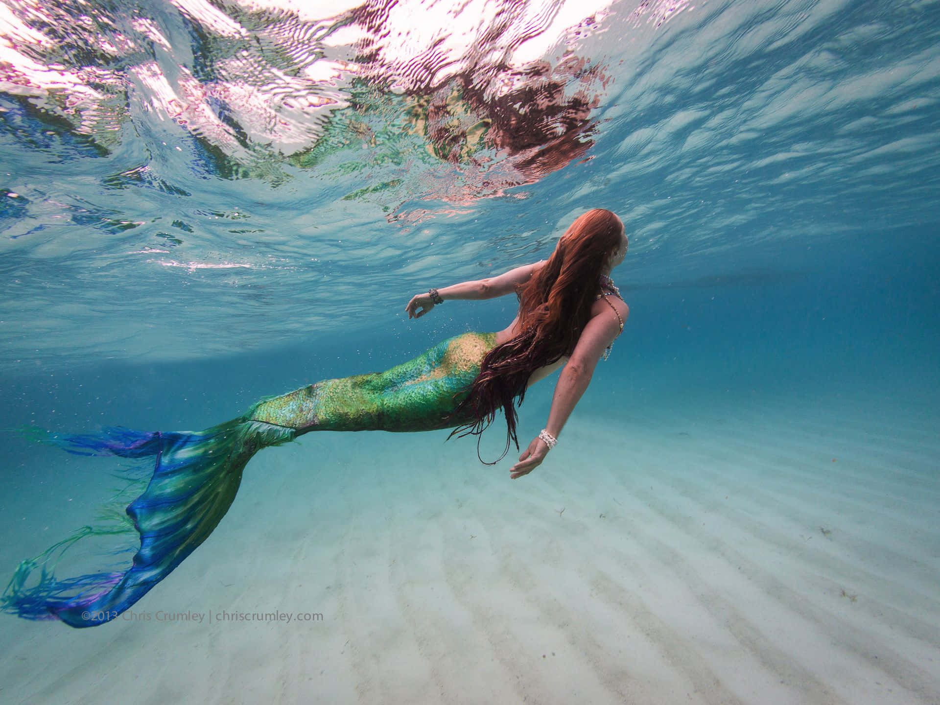 Real Mermaid 桌布 壁紙 — 一條長著紅色長髮的美人魚在波光粼粼的陽光下，優雅地在沙質海床上滑行。