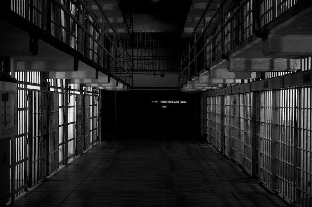 Prison 桌布 壁紙 — 一條長長的、燈光昏暗的監獄走廊，兩邊都有鐵欄牢房，通向黑暗深處。