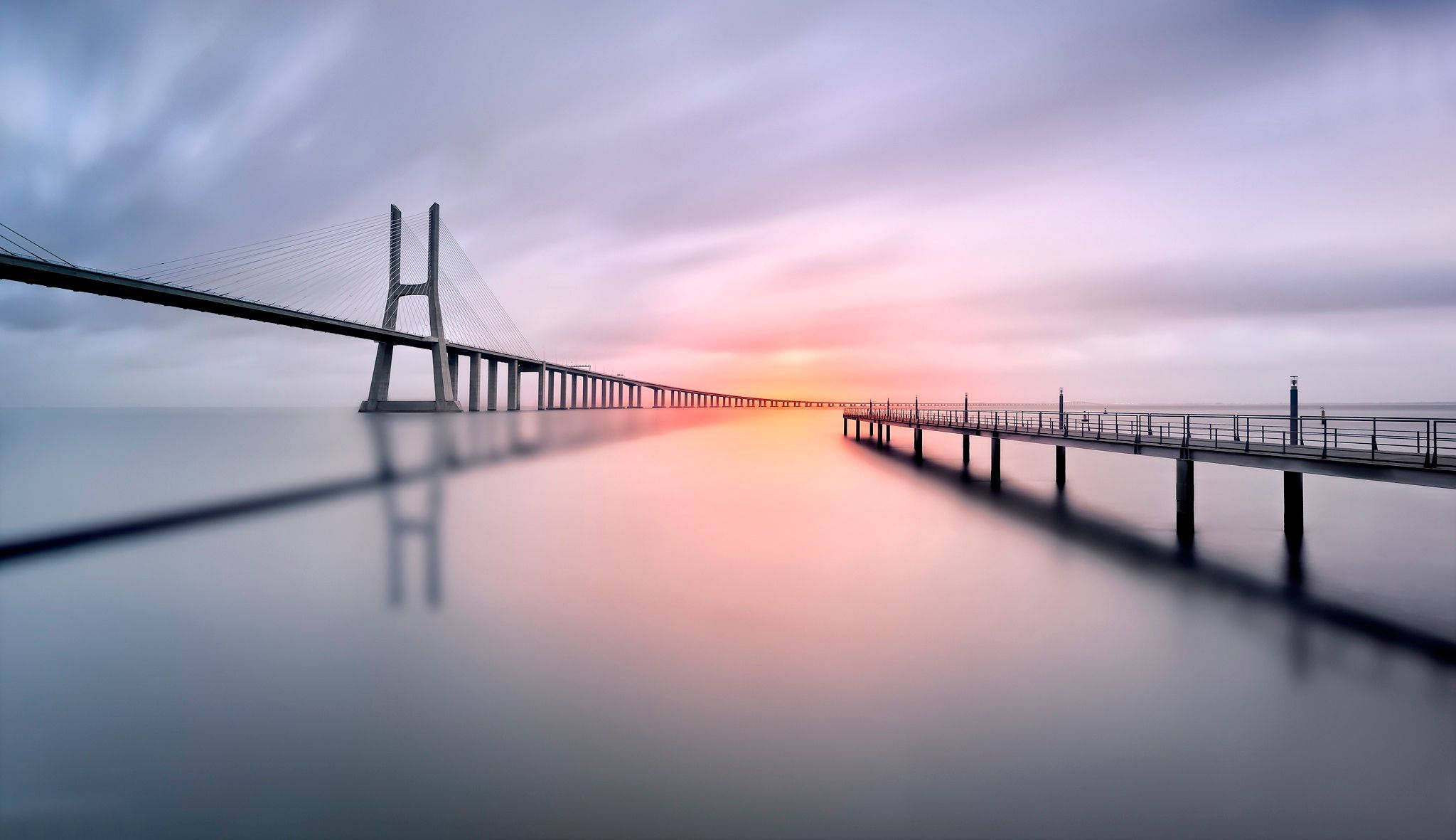 Minimalist Nature 桌布 壁紙 — 一條長長的吊橋和碼頭在柔和粉紅和紫色的日落天空下，延伸到平靜的水面上。