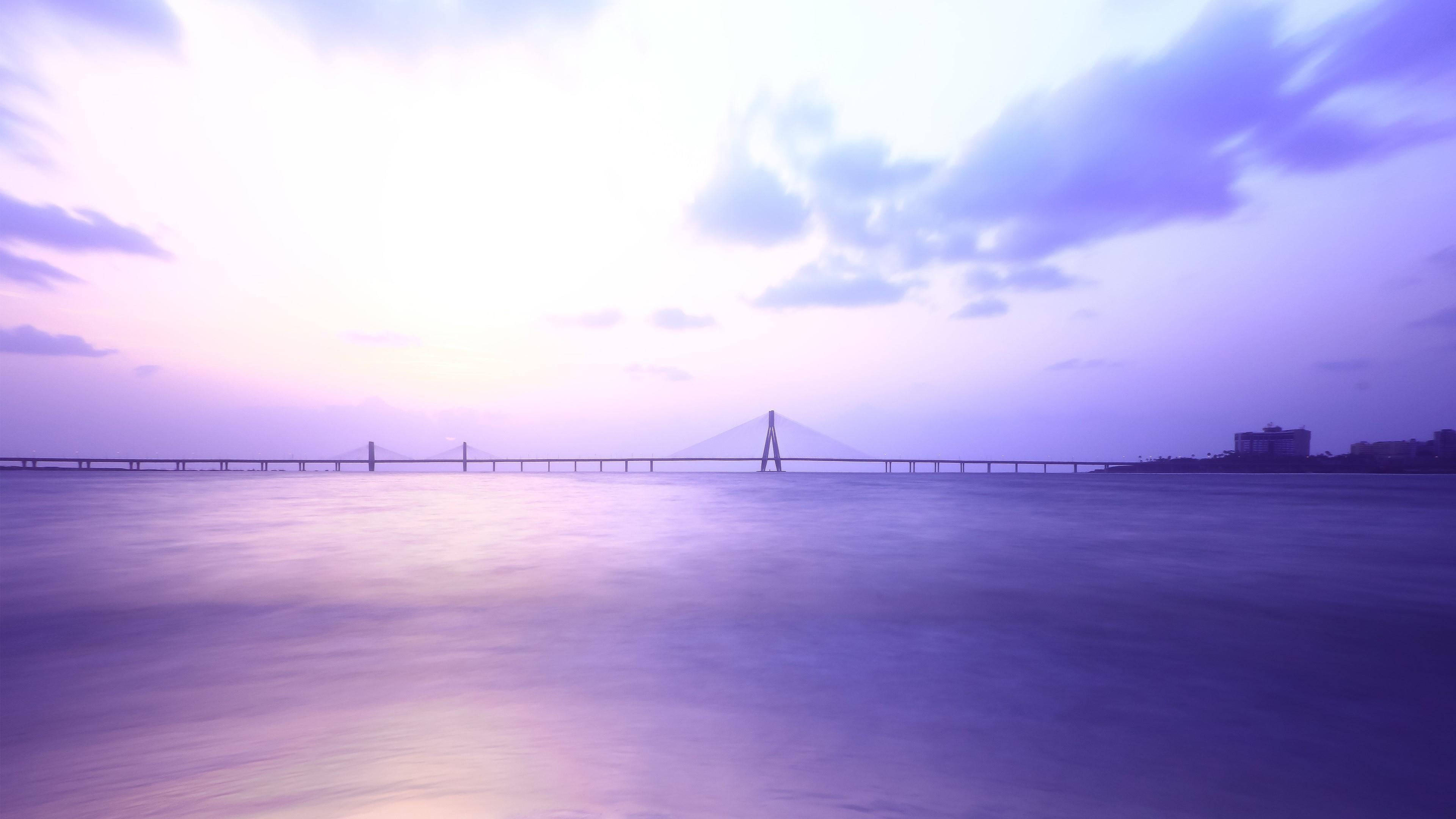Mumbai 桌布 壁紙 — 一條長長的吊橋橫跨平靜的水面，上方是柔和的紫色和粉紅色暮色天空，雲朵模糊。