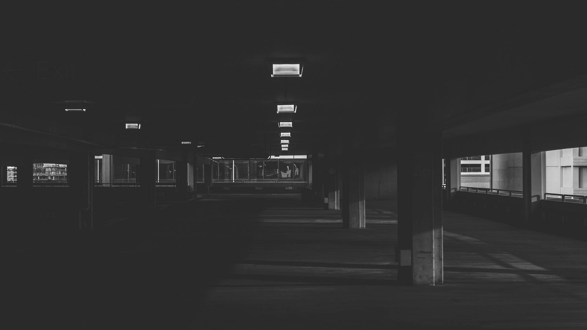 Monochrome 桌布 壁紙 — 一條長長的昏暗地下通道，有混凝土柱和遠處發光的出口標誌，投射出鮮明的陰影。