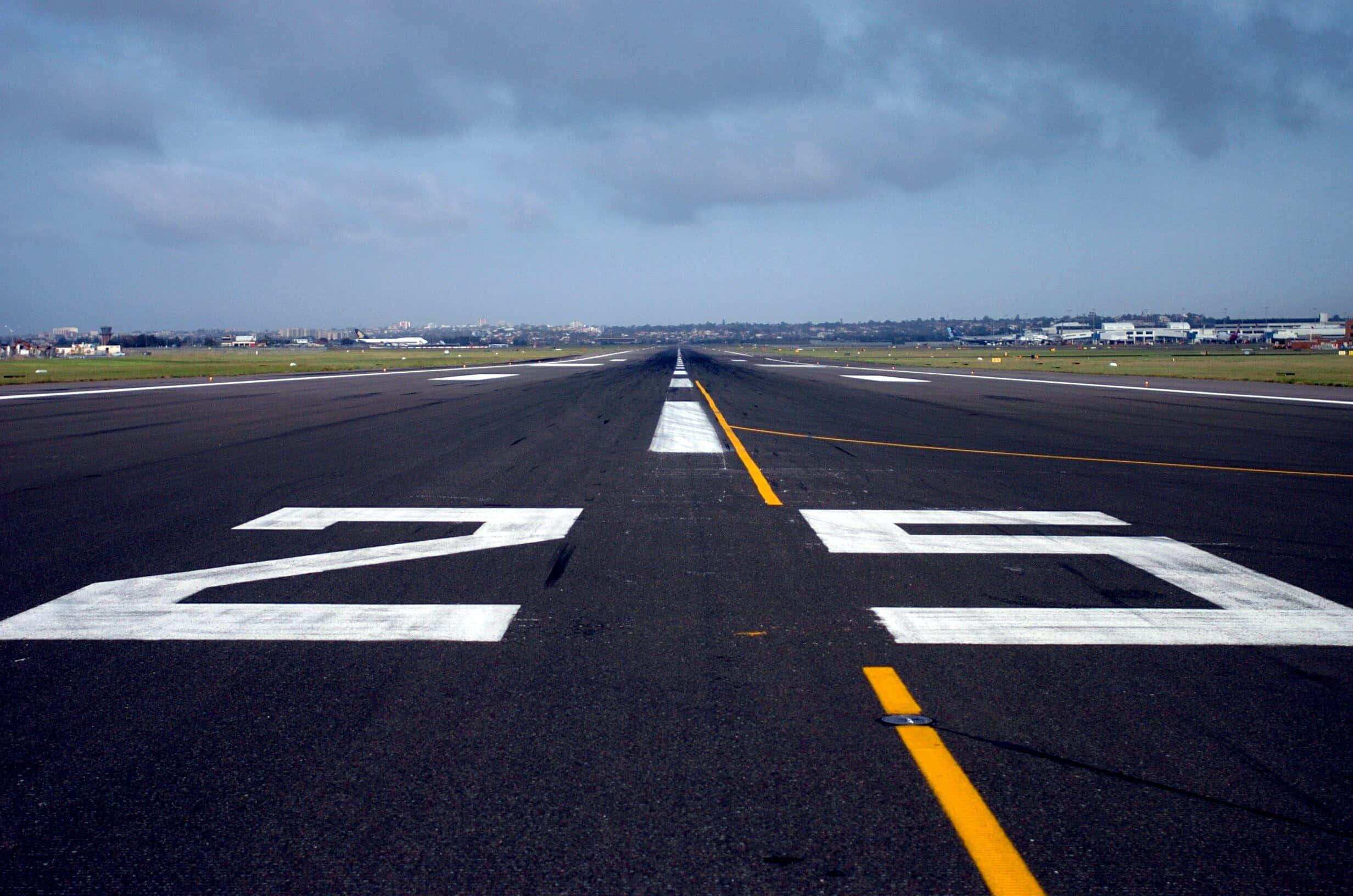 Runway 桌布 壁紙 — 一條長長的機場跑道伸向多雲的地平線，在陰天之下帶有醒目的白色和黃色標記。