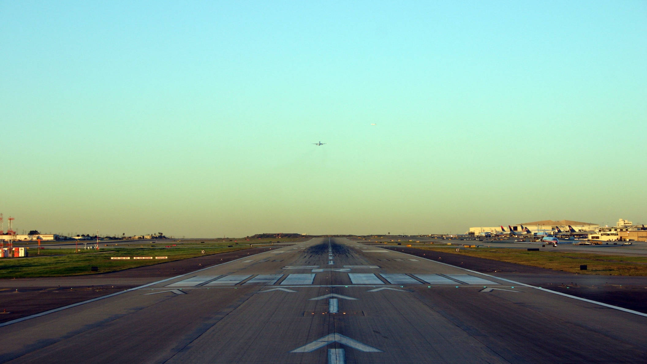 Runway 桌布 壁紙 — 一條長長的機場跑道向遠方地平線延伸，一架小型飛機在黃金時段從頭頂飛過。