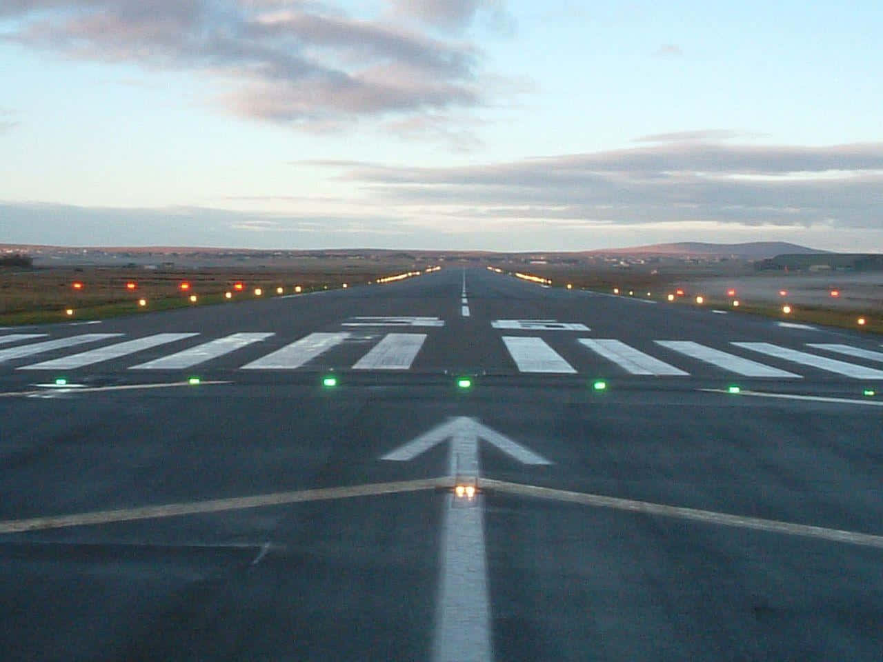 Runway 桌布 壁紙 — 一條長長的機場跑道在柔和的暮色天空下延伸至地平線，跑道邊緣有發光的燈光，停機坪上畫有一個大箭頭。