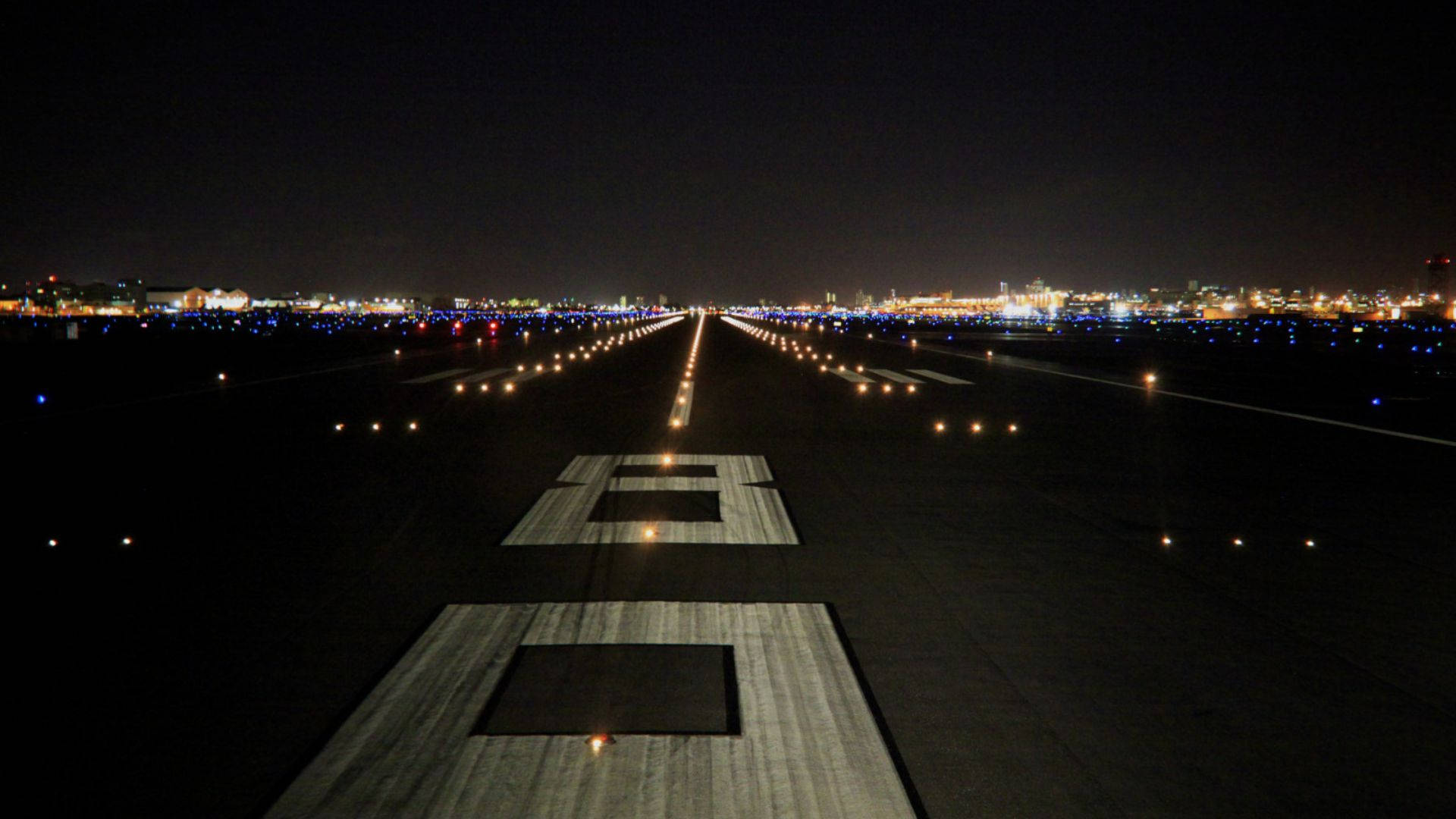 Runway 桌布 壁紙 — 一條長長的機場跑道延伸至夜晚，被發光的燈光照亮，地平線上有遠處的城市燈光。
