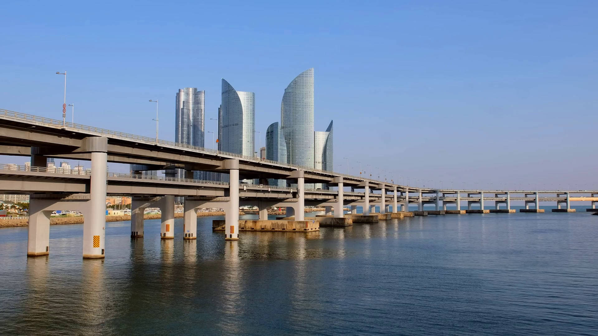 Busan 桌布 壁紙 — 一條長長的混凝土橋樑橫跨平靜的水面，通往未來主義的玻璃摩天大樓，在清澈的藍天下。