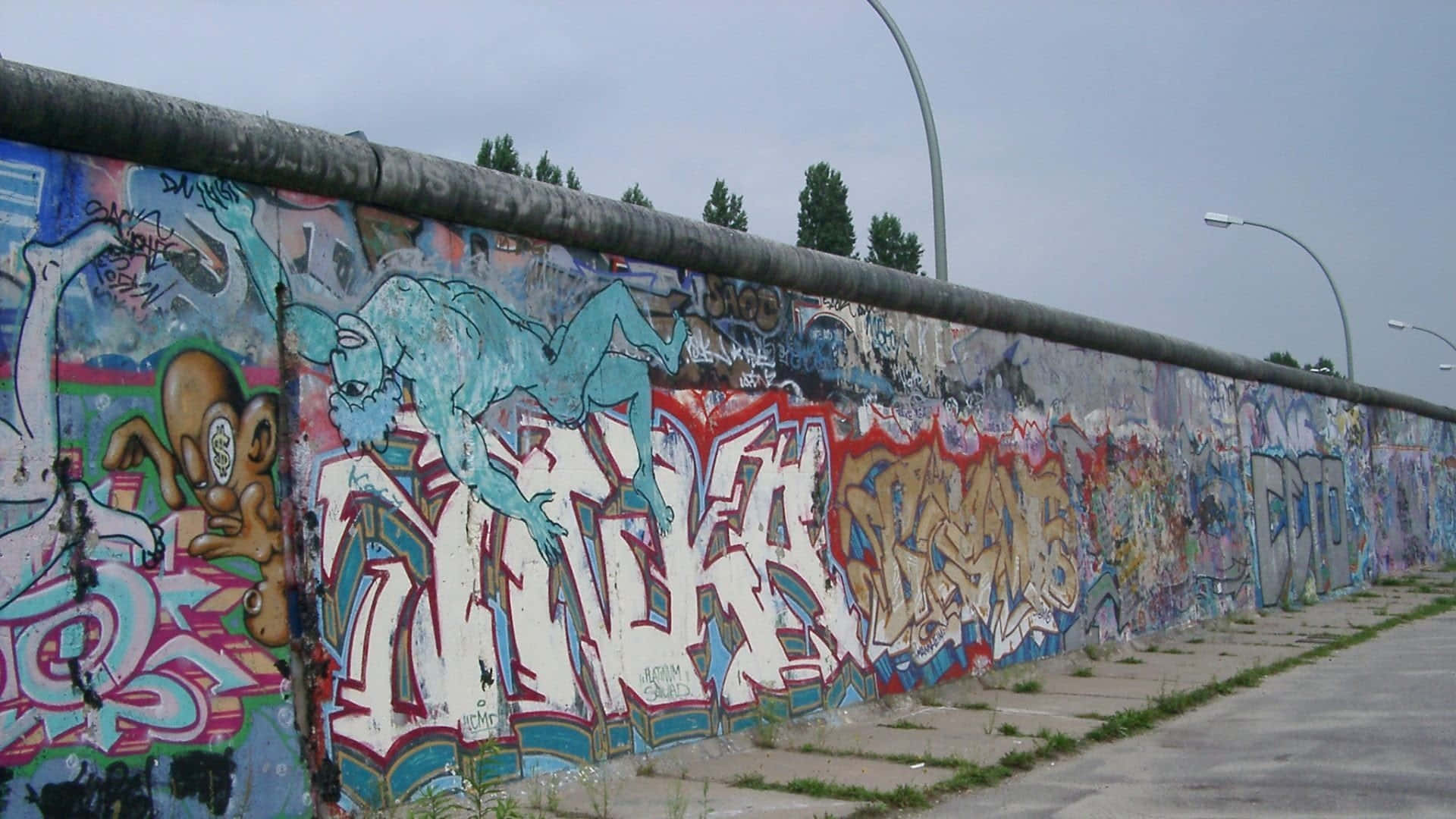 wall 桌布 壁紙 — 一條長長的混凝土牆，在陰天背景下，覆蓋著充滿活力的塗鴉藝術，背景有街燈和樹木。
