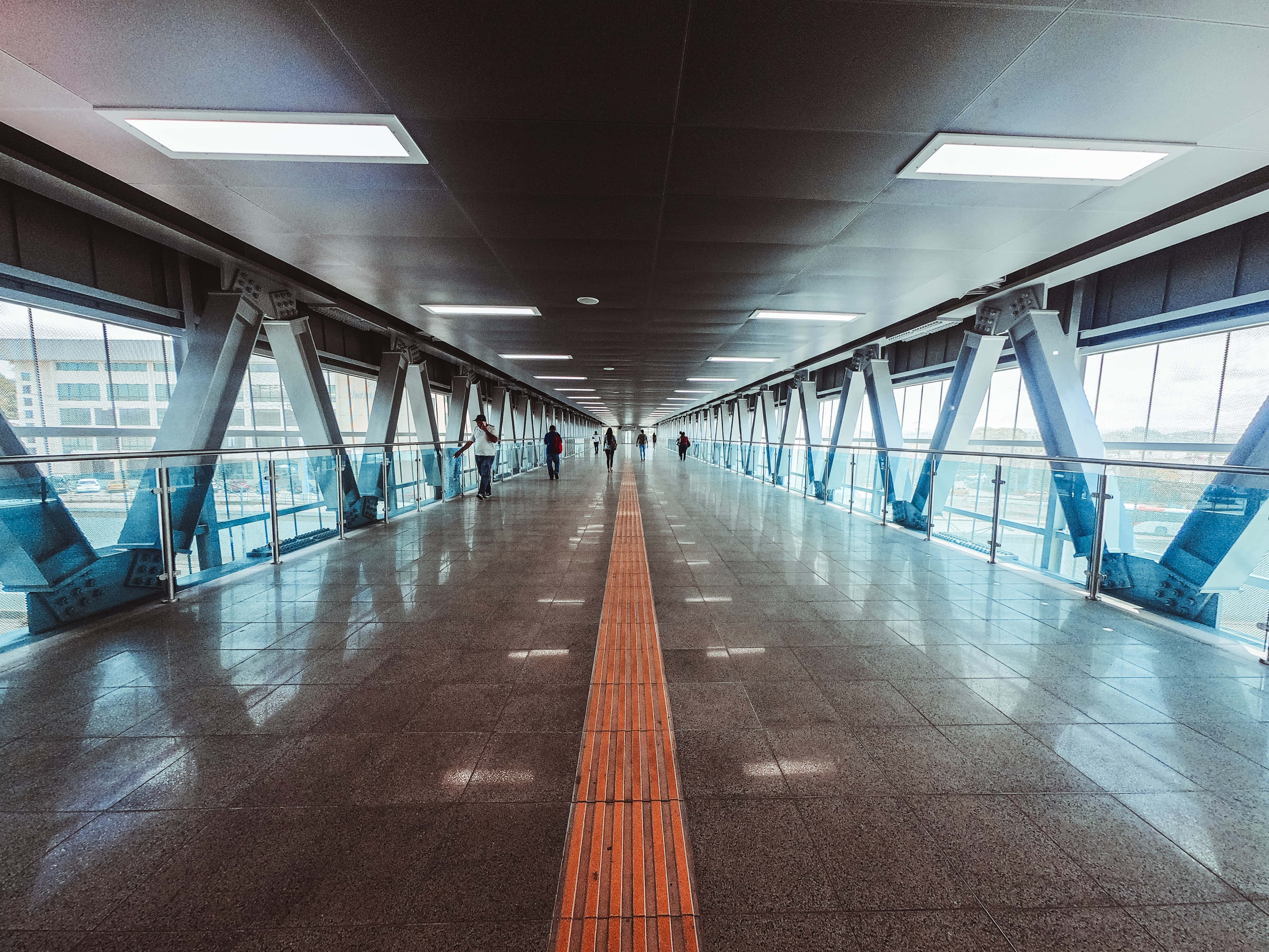 機場 桌布 壁紙 — 一條長長的現代行人天橋，帶有反光地板和藍色結構支撐，在明亮的頭頂燈光下延伸至遠方。