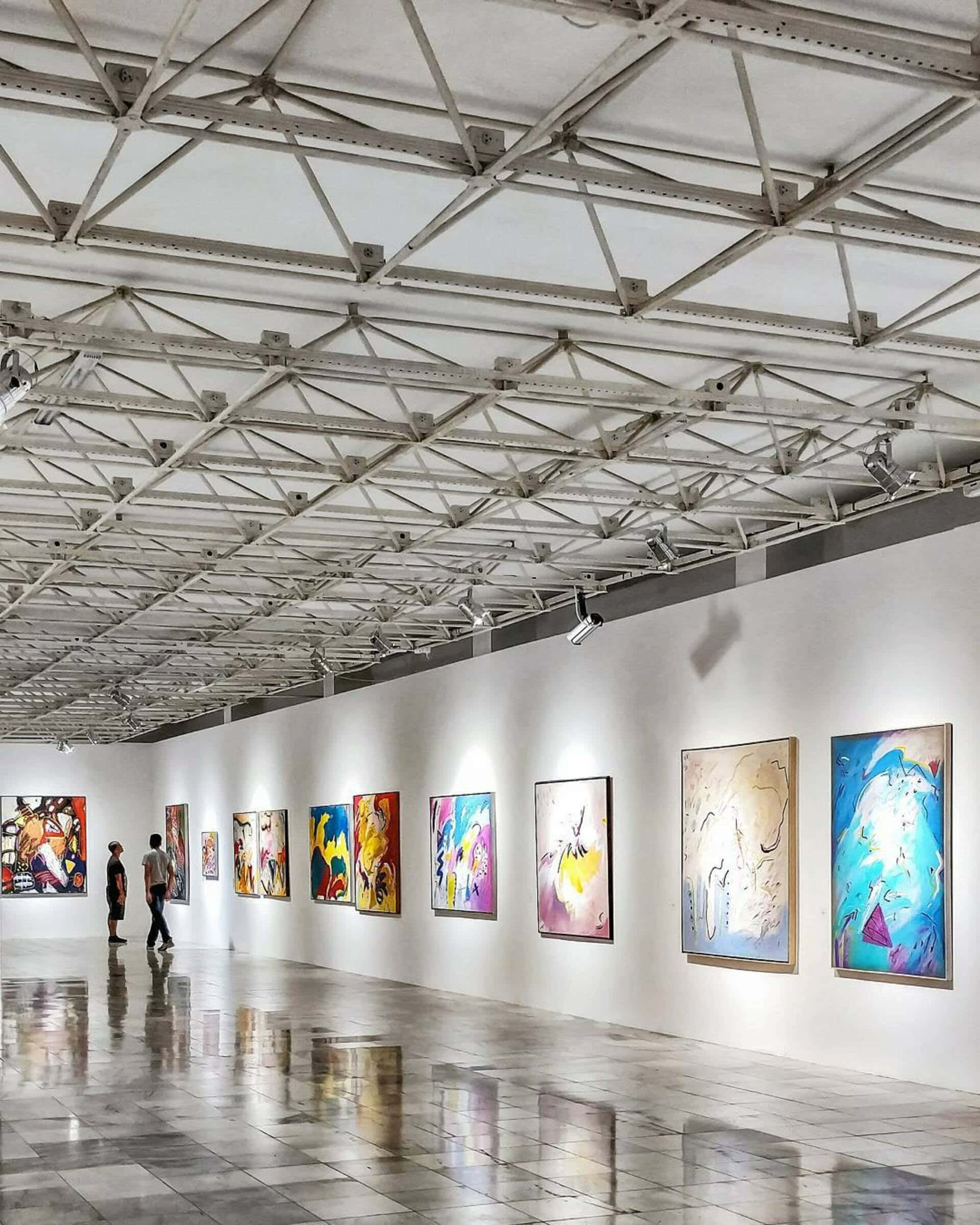 Gallery 桌布 壁紙 — 一條長長的畫廊走廊，白色牆壁上展示著充滿活力的抽象畫，下方是工業風格的屋頂桁架和聚光燈。