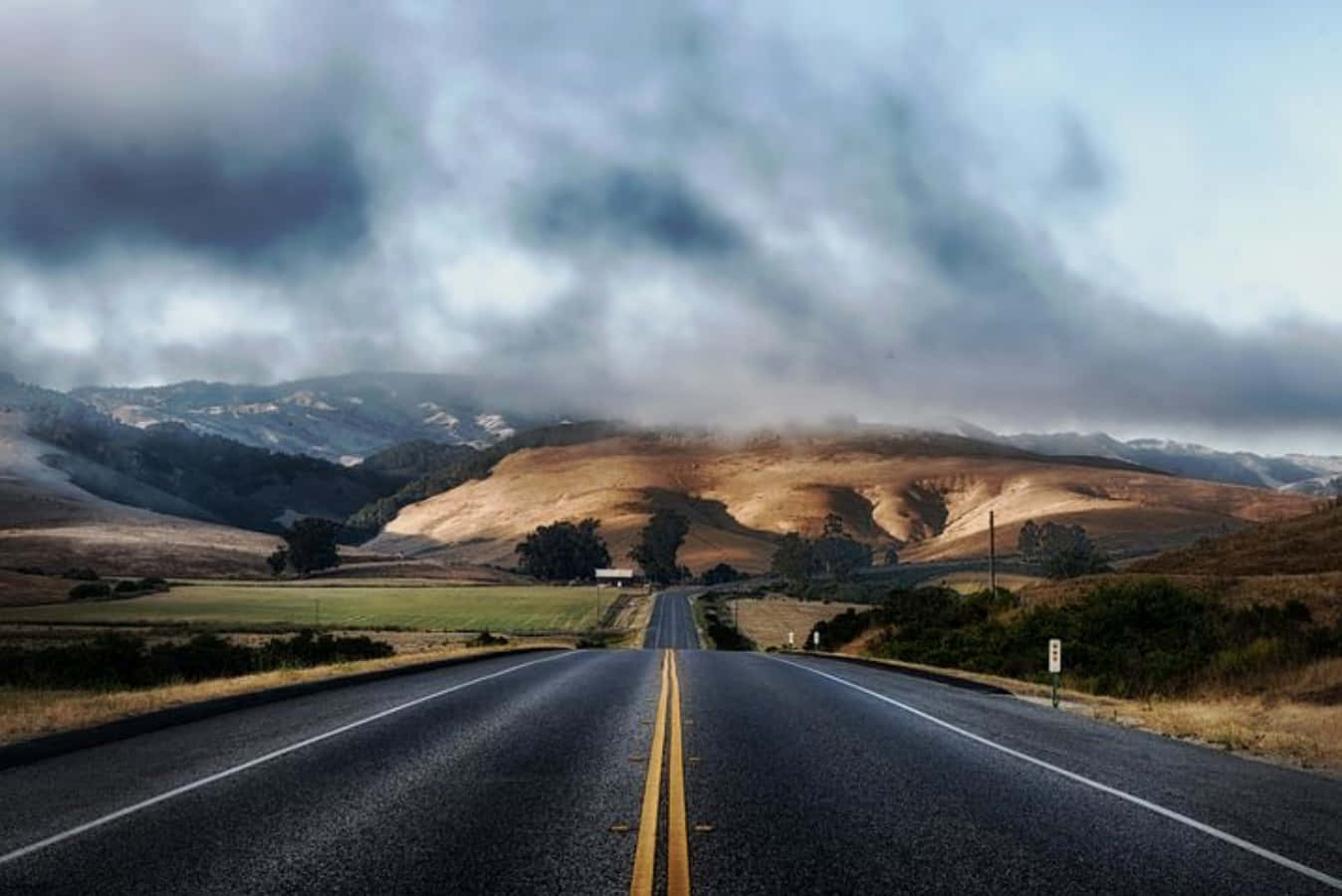 Highways 桌布 壁紙 — 一條長長的直路在戲劇性的多雲天空下延伸至遠山，兩旁是金色的田野。