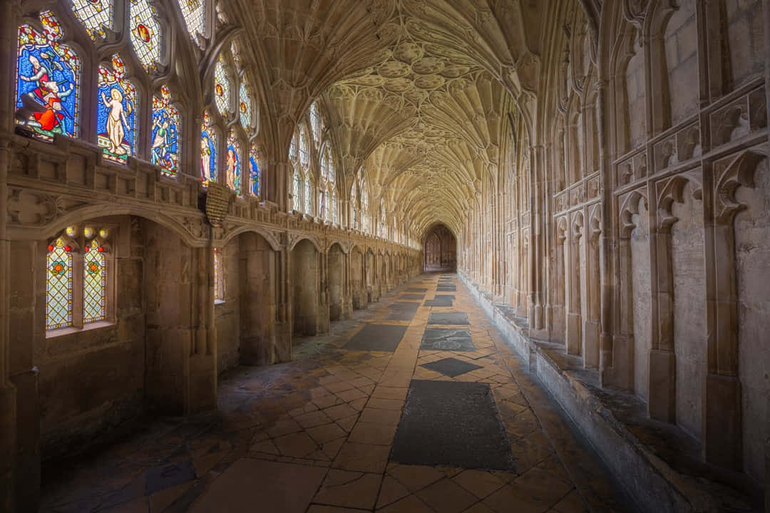 The Hogwarts Charms Corridor 桌布 壁紙 — 一條長長的石頭迴廊，擁有拱形天花板和彩色玻璃窗，沐浴在柔和自然光中。