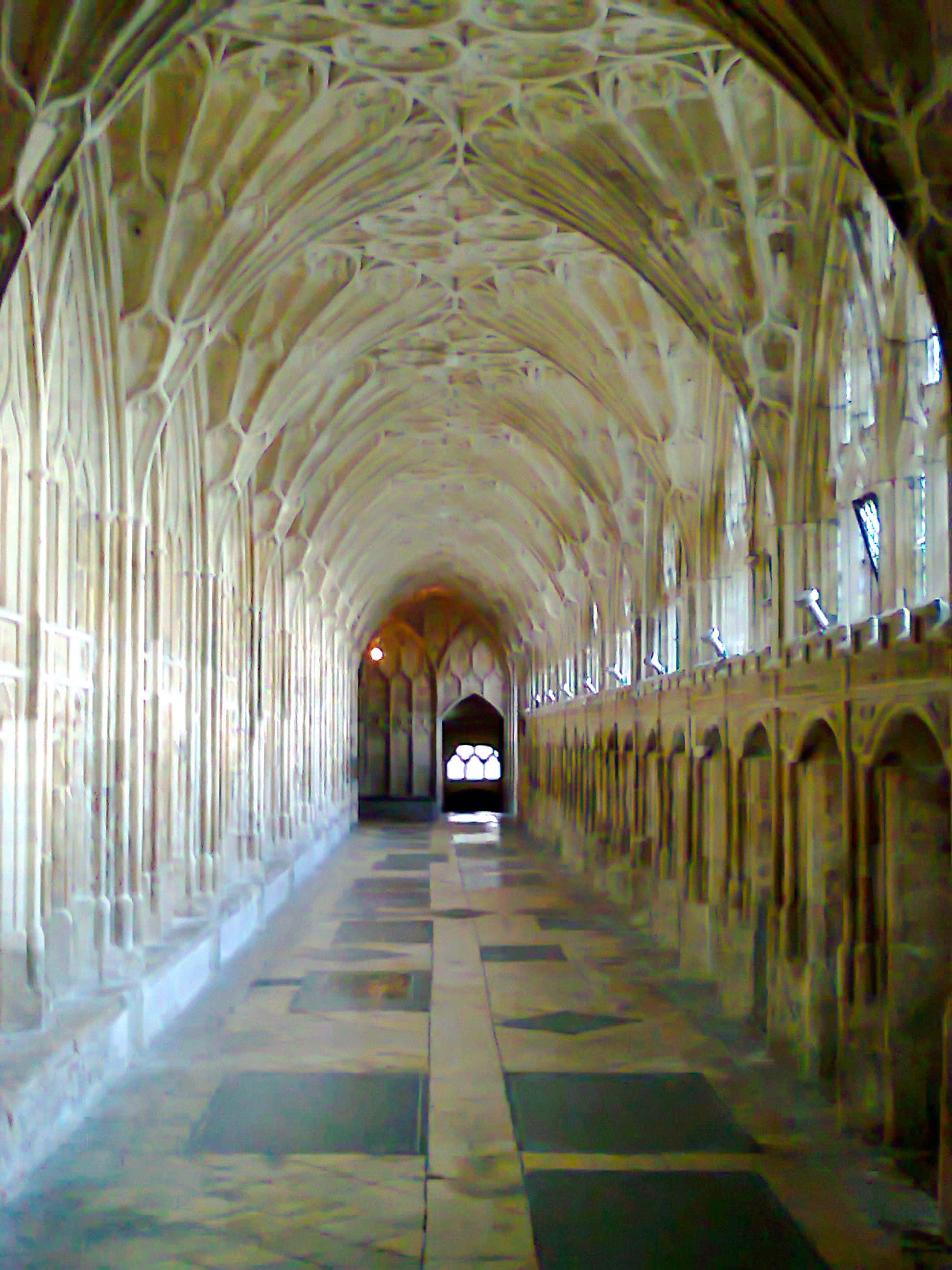 The Hogwarts Charms Corridor 桌布 壁紙 — 一條長長的石頭迴廊，擁有精緻的拱形天花板和拱形窗戶，沐浴在柔和自然光中。