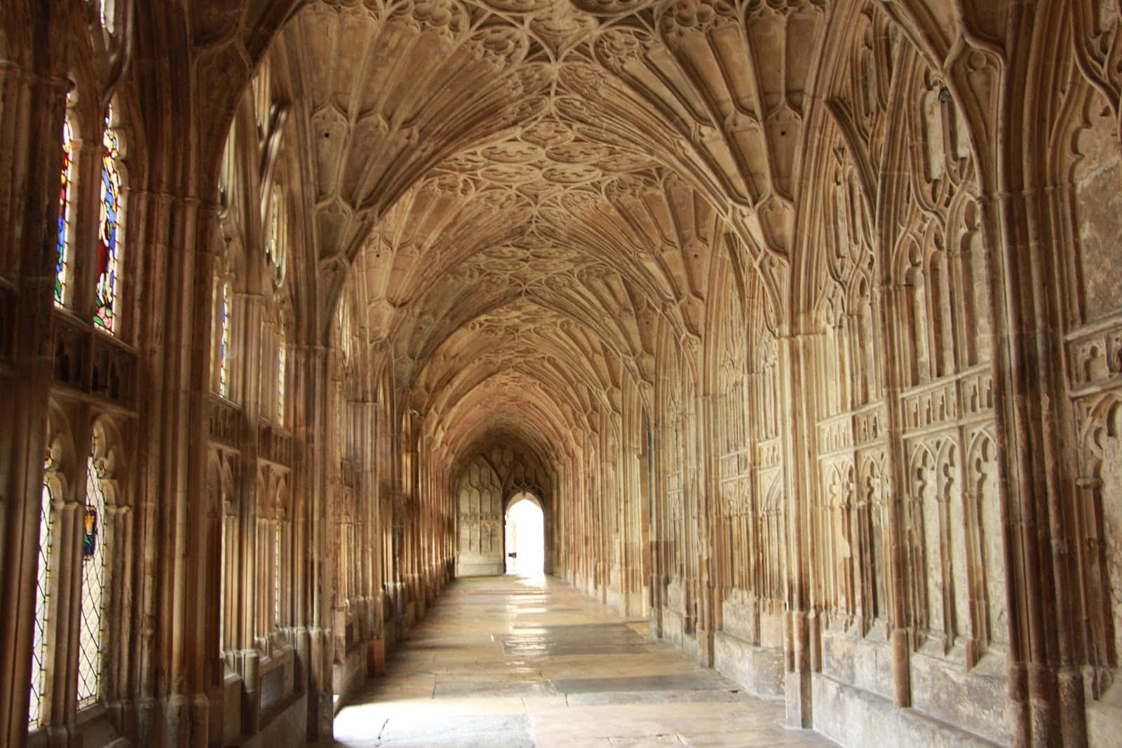 The Hogwarts Charms Corridor 桌布 壁紙 — 一條長長的石頭迴廊，擁有精緻的拱形天花板和彩色玻璃窗，沐浴在溫暖的陽光中。