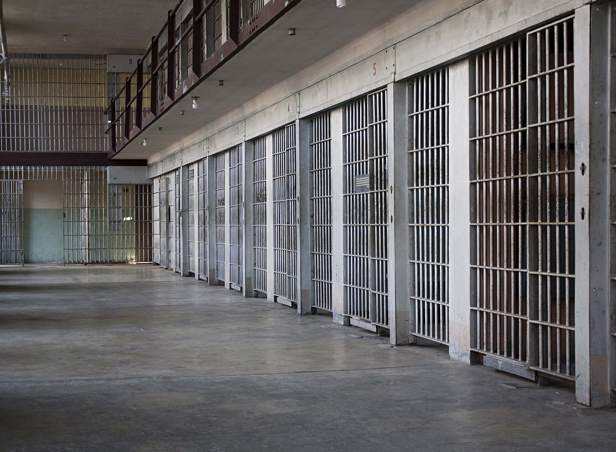 Prison 桌布 壁紙 — 一條長長的空蕩監獄走廊，兩排鐵欄牢房和二樓通道，在昏暗的螢光燈下。