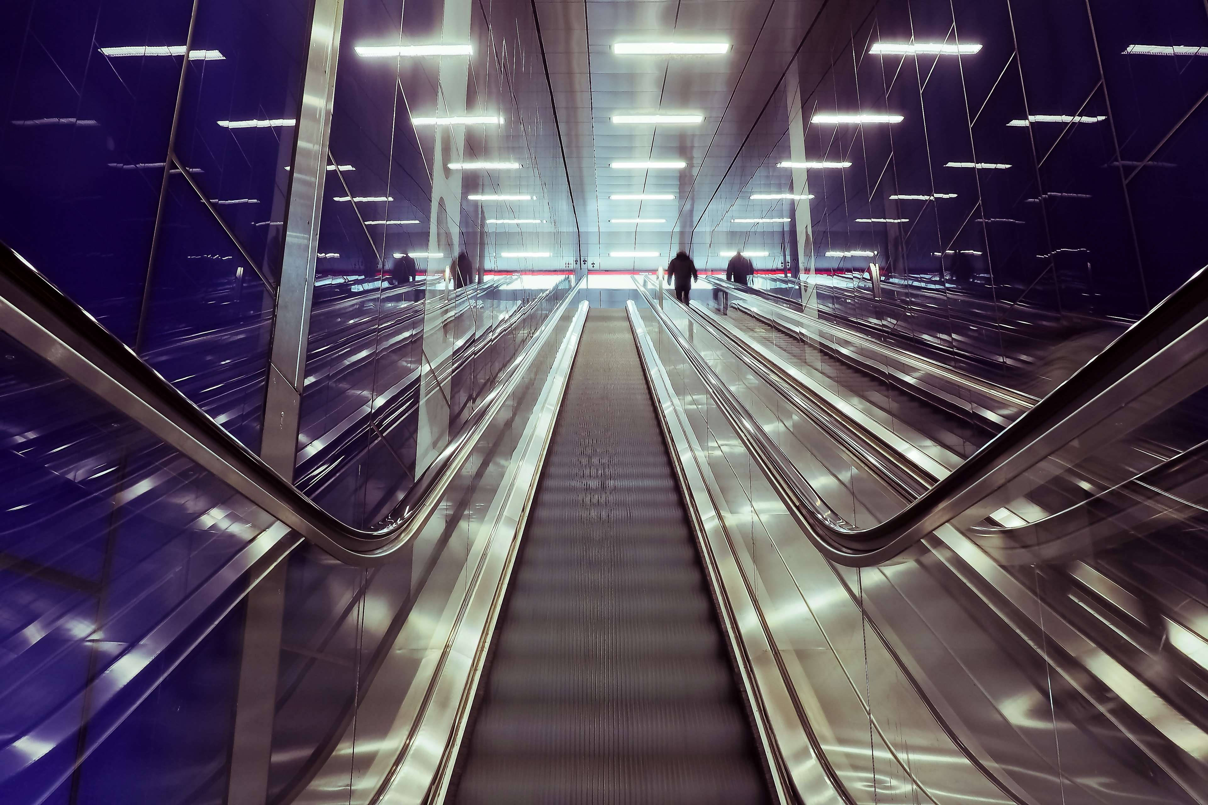 Airport 桌布 壁紙 — 一條長長的自動扶梯隧道，帶有反光玻璃牆和發光的頭頂燈光，向上通往遠處的通勤者。