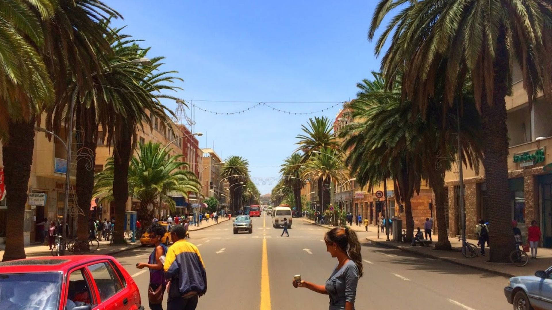 Eritrea 桌布 壁紙 — 一條陽光明媚的城市街道，兩旁種滿高大的棕櫚樹，行人走過色彩繽紛的建築物，天空湛藍清澈。